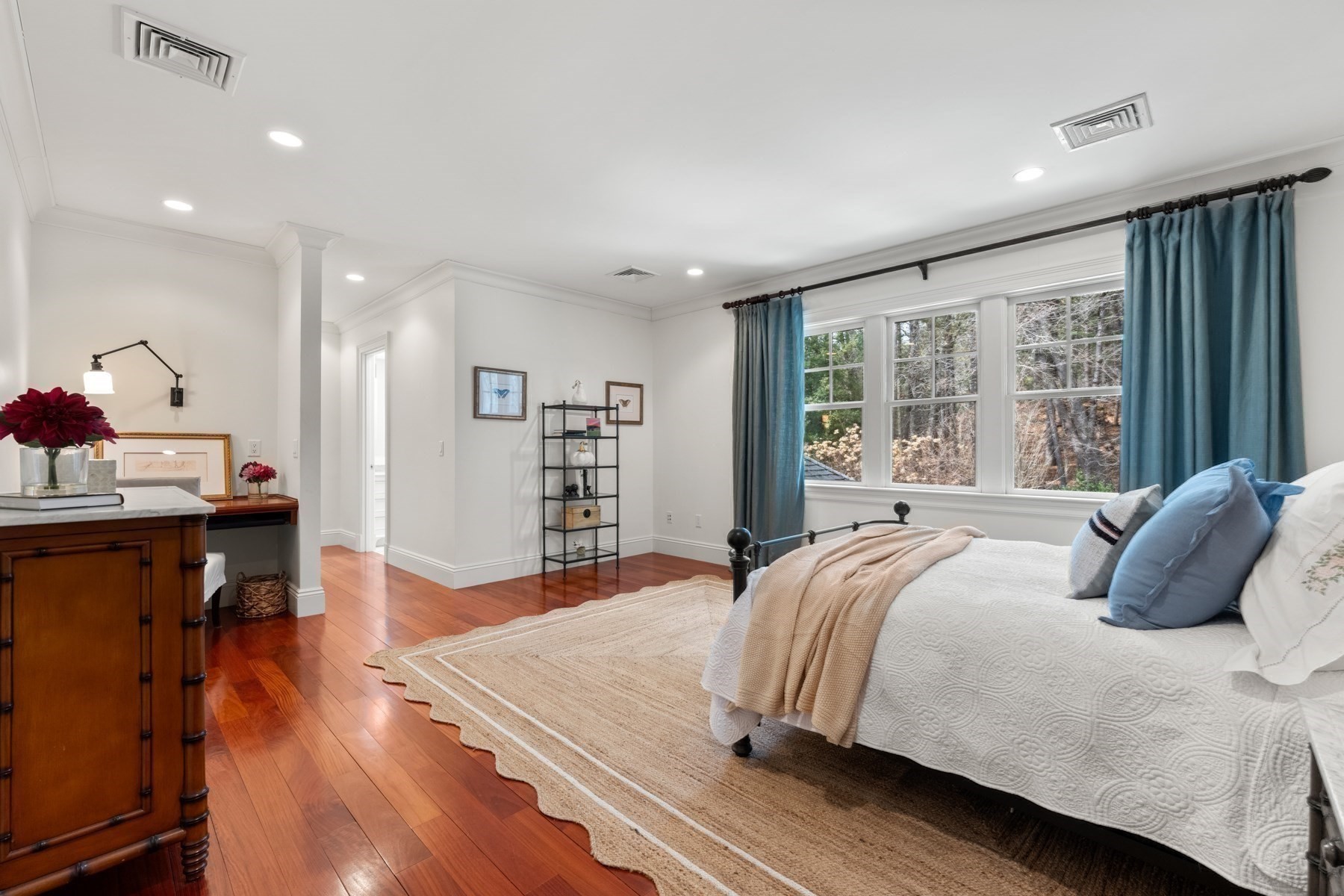 Bedroom with a white quilted bed, blue curtains, warm hardwood floors, and large windows overlooking trees.