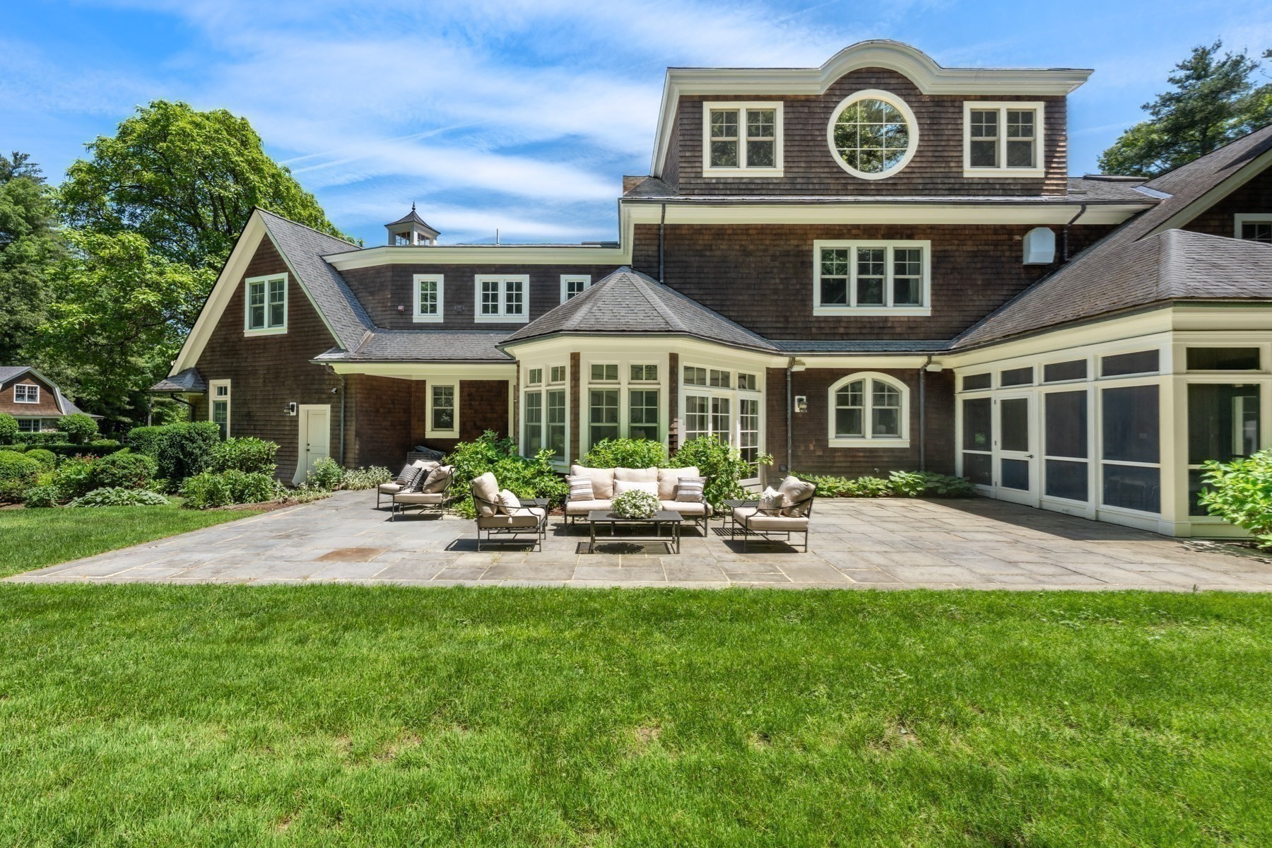 Rear of a shingle home with a round upper window, glass sunroom, and furnished stone patio.