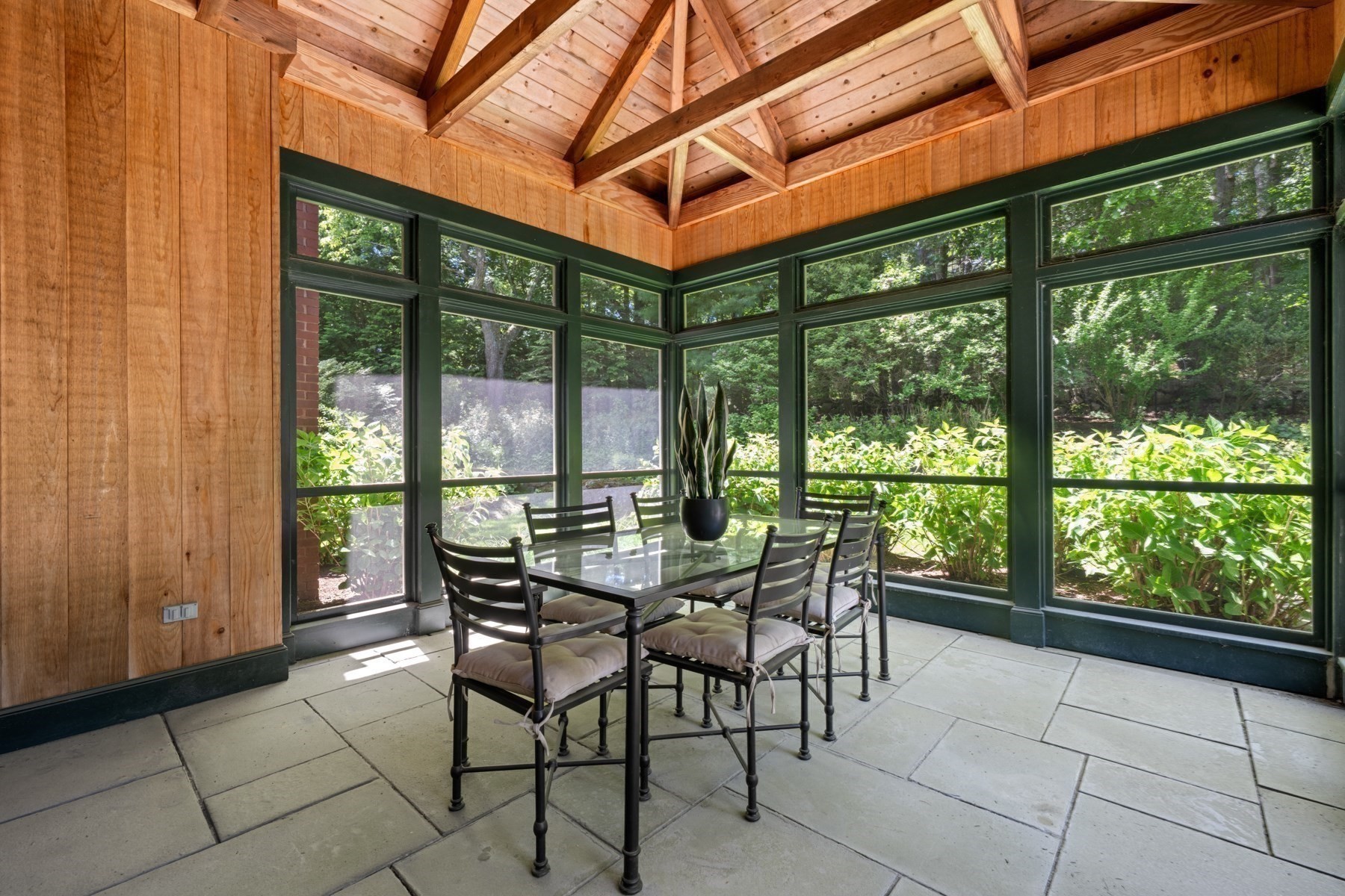 Sunroom with a glass dining table, leafy garden views, and an exposed vaulted wood ceiling.