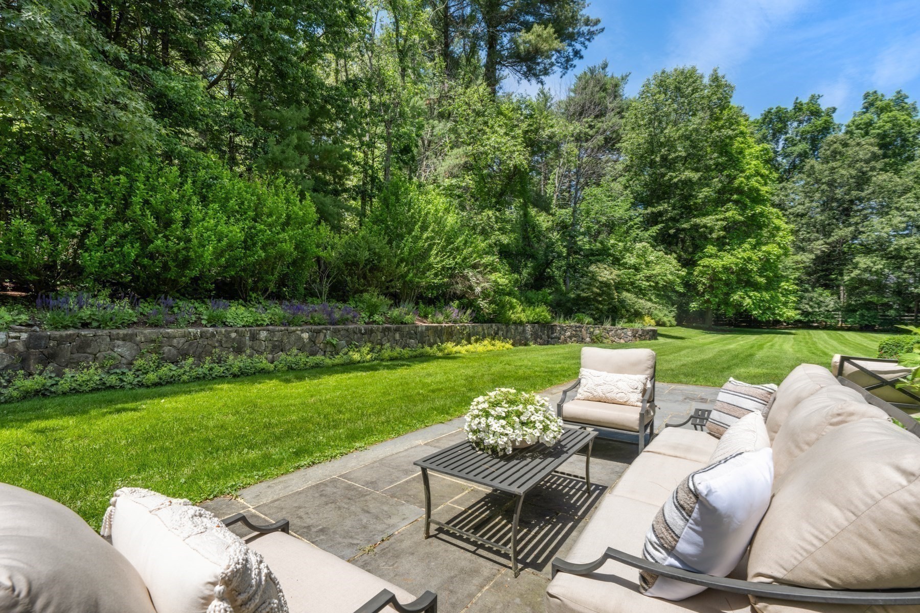 Stone patio with pale outdoor seating overlooking a lawn and mature trees.