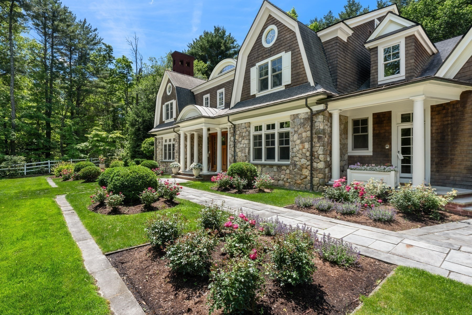 Shingle-style house with a stone facade, white columns, and flowering garden along a paved walkway.