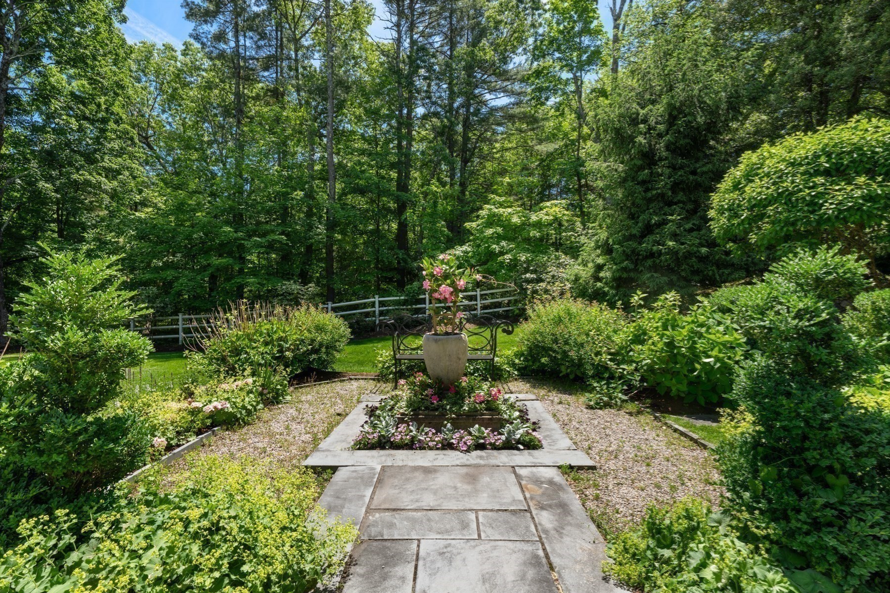Landscaped garden with a stone path, flower bed, urn, and bench beneath mature trees.