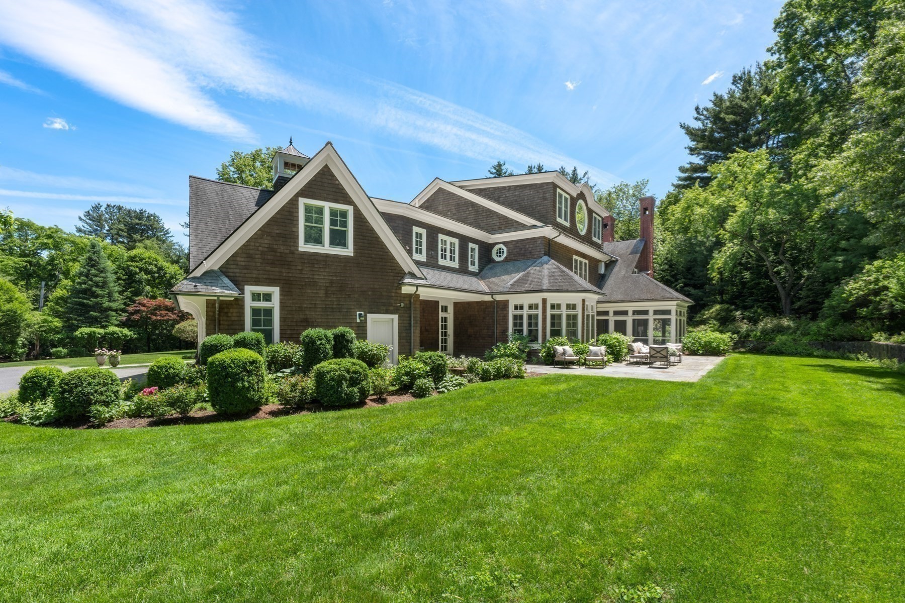 Brown shingle house with a glass sunroom, stone patio, landscaped lawn, and mature trees.