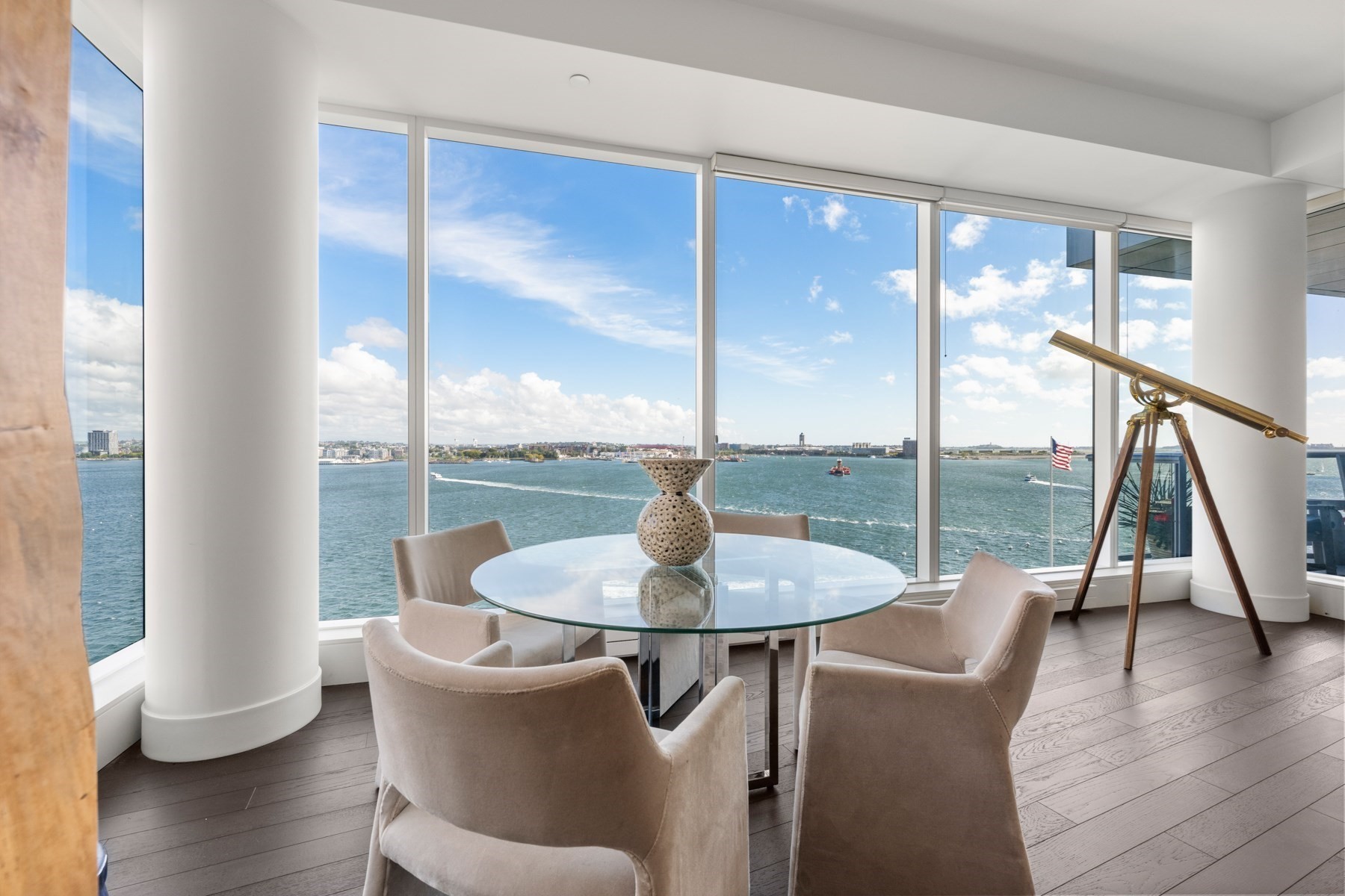 Dining area with a glass table and brass telescope overlooking the water through expansive windows.