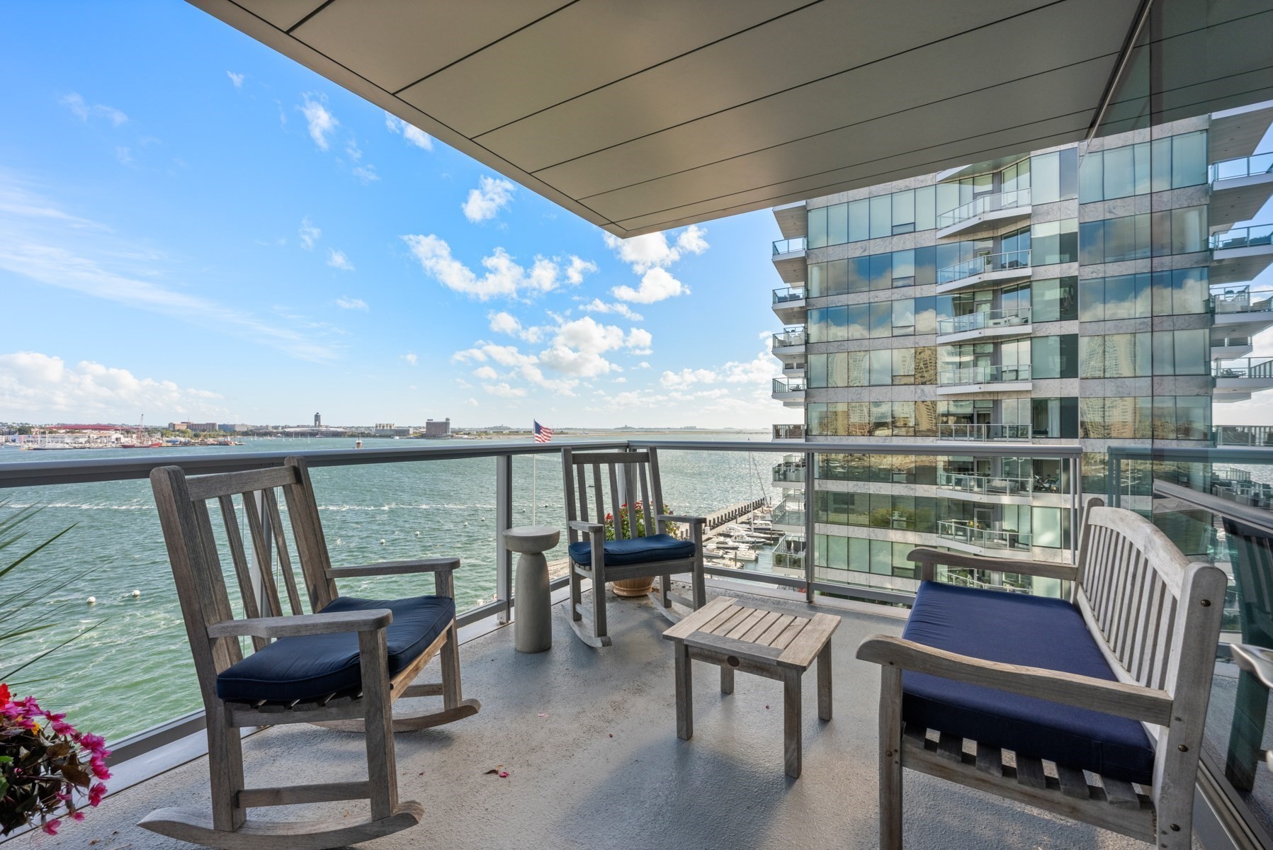 Waterfront balcony with cushioned seating overlooking the harbor and a neighboring glass apartment building.
