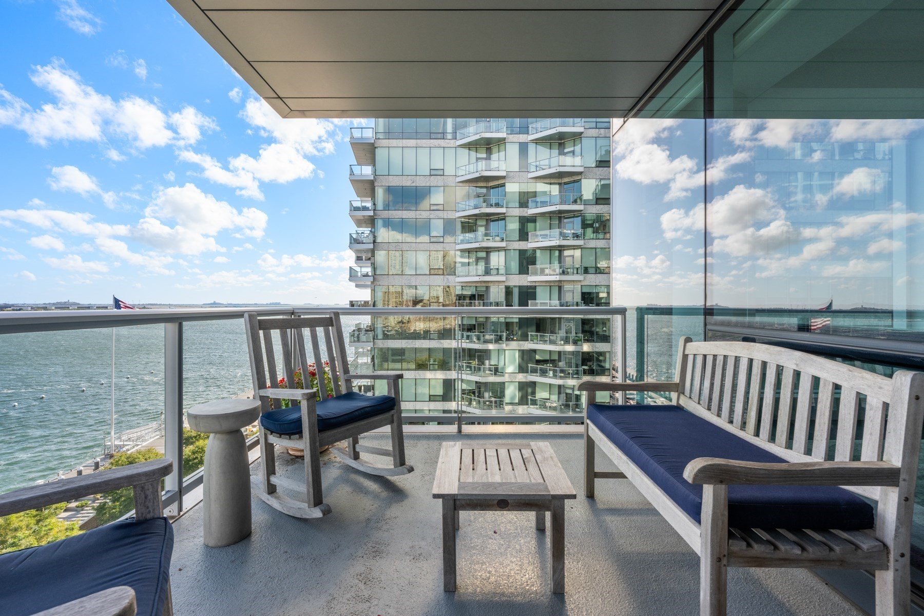 Furnished high-rise balcony overlooking harbor water and a modern glass apartment building.