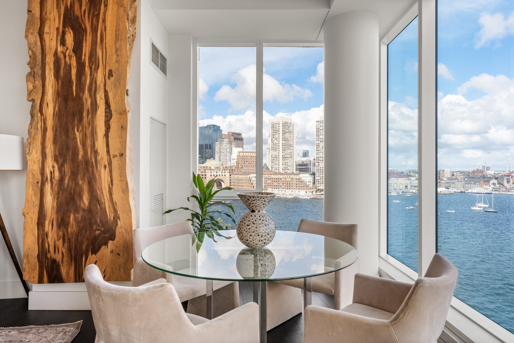 Dining area with a round glass table, harbor views, and Boston skyline through floor-to-ceiling windows.