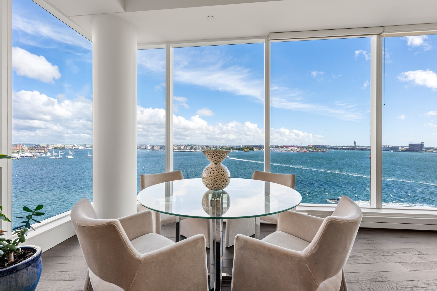 Dining area with floor-to-ceiling harbor views and a round glass table surrounded by upholstered chairs.