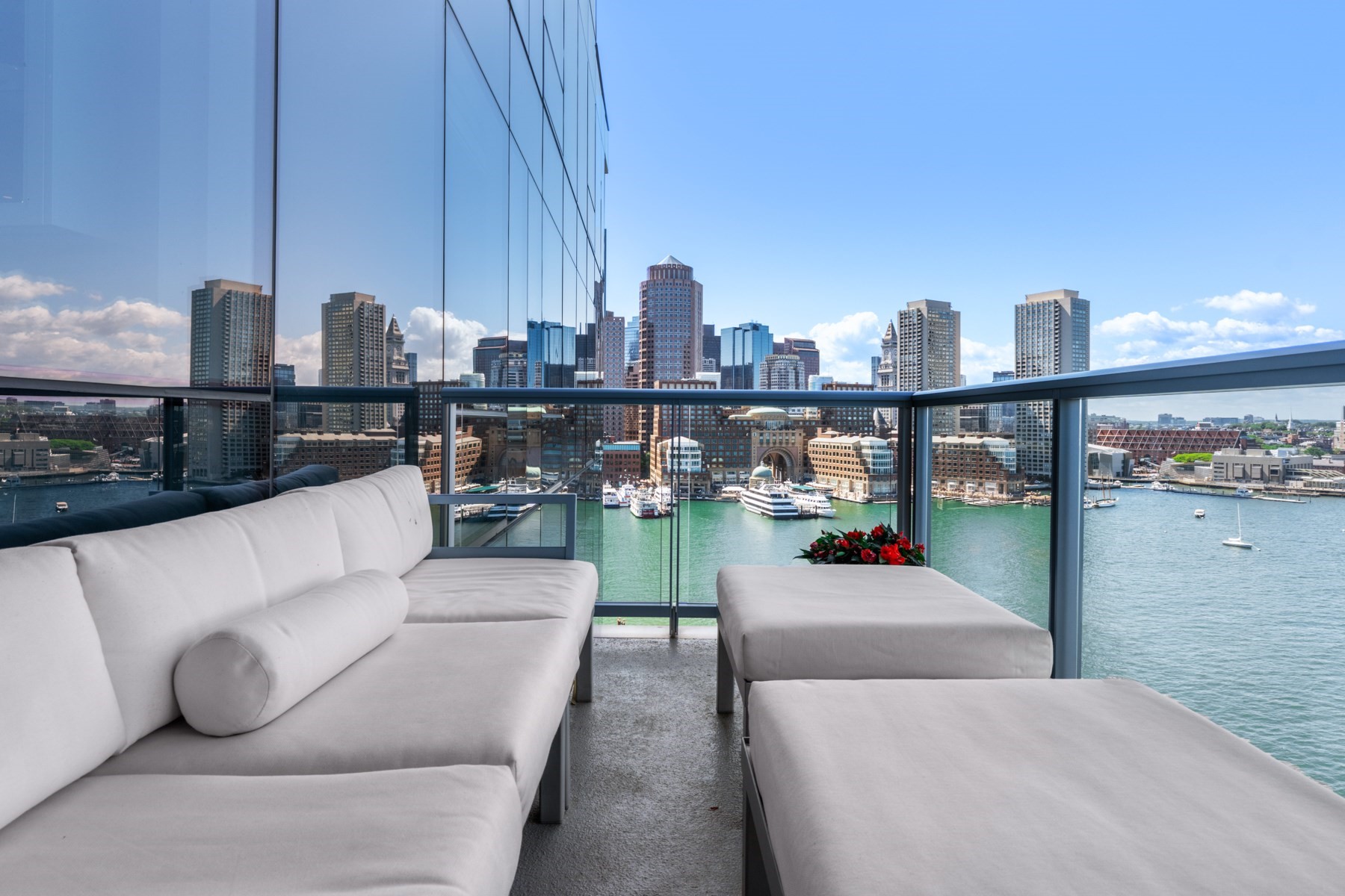 Furnished balcony overlooking Boston Harbor and the downtown skyline, with white seating and glass railings.