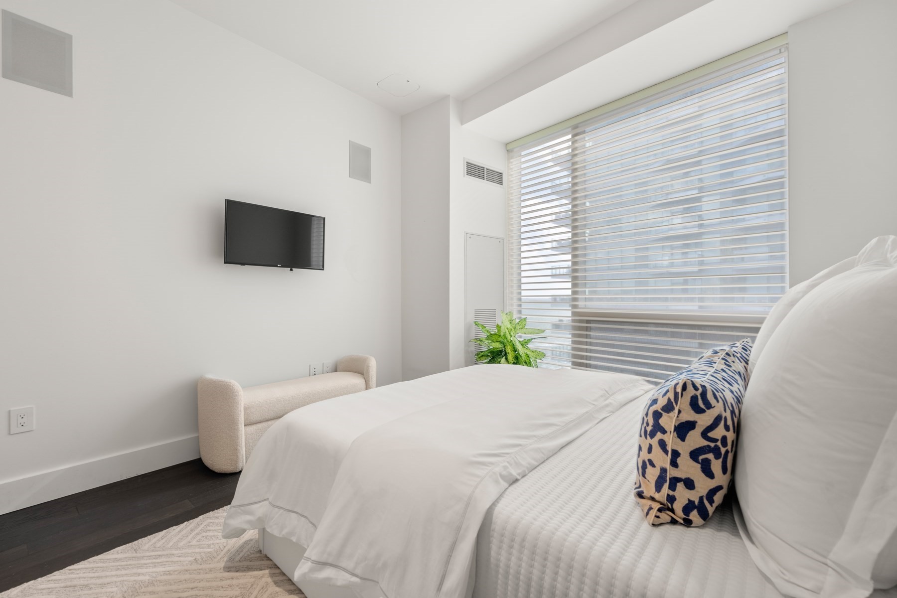 Bedroom with a white bed, patterned accent pillow, and large window blinds overlooking a neighboring building.