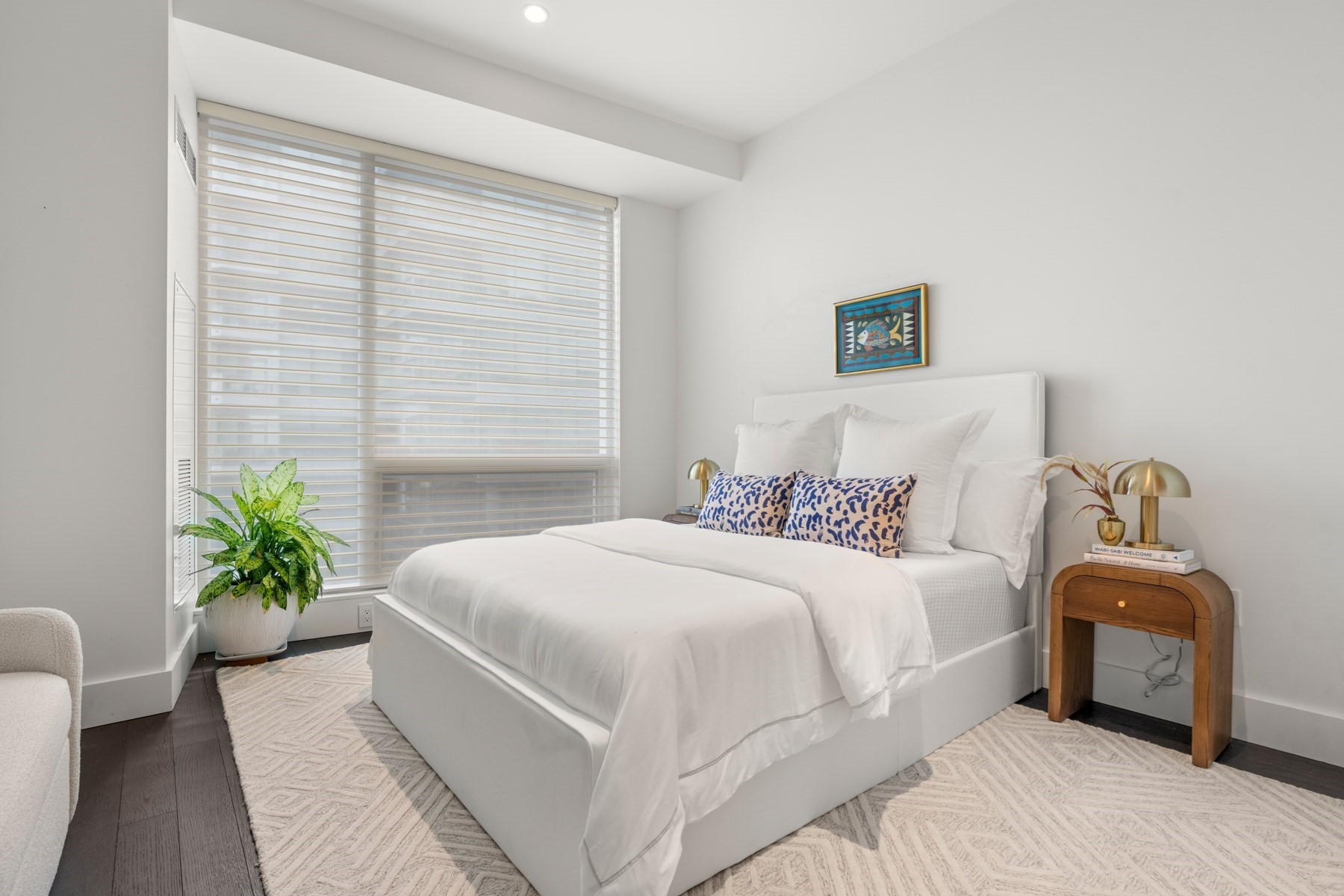 Bedroom with a white bed, blue patterned pillows, large shaded windows, and a potted plant.