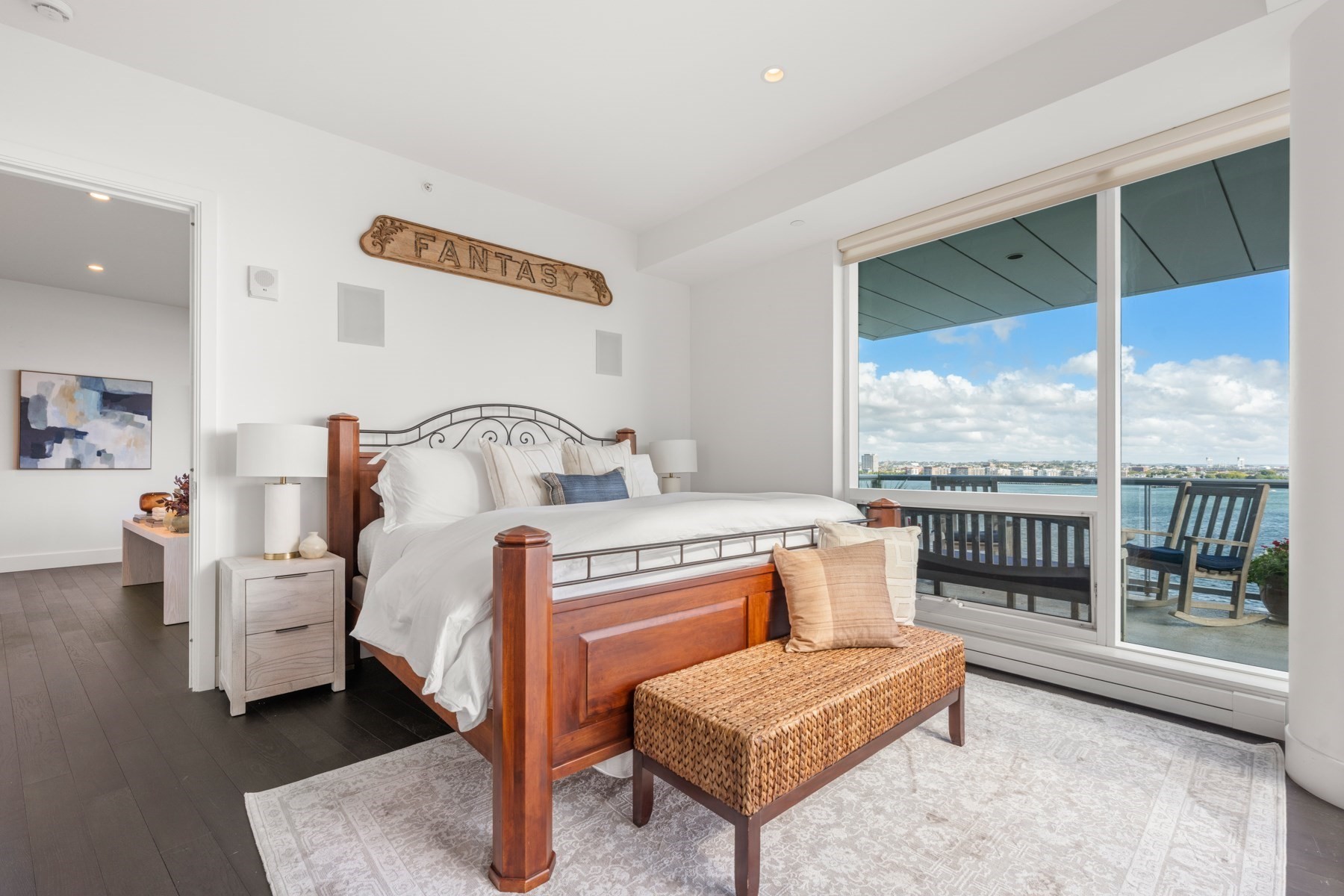 Bedroom with a wooden bed and balcony overlooking the water and distant buildings.