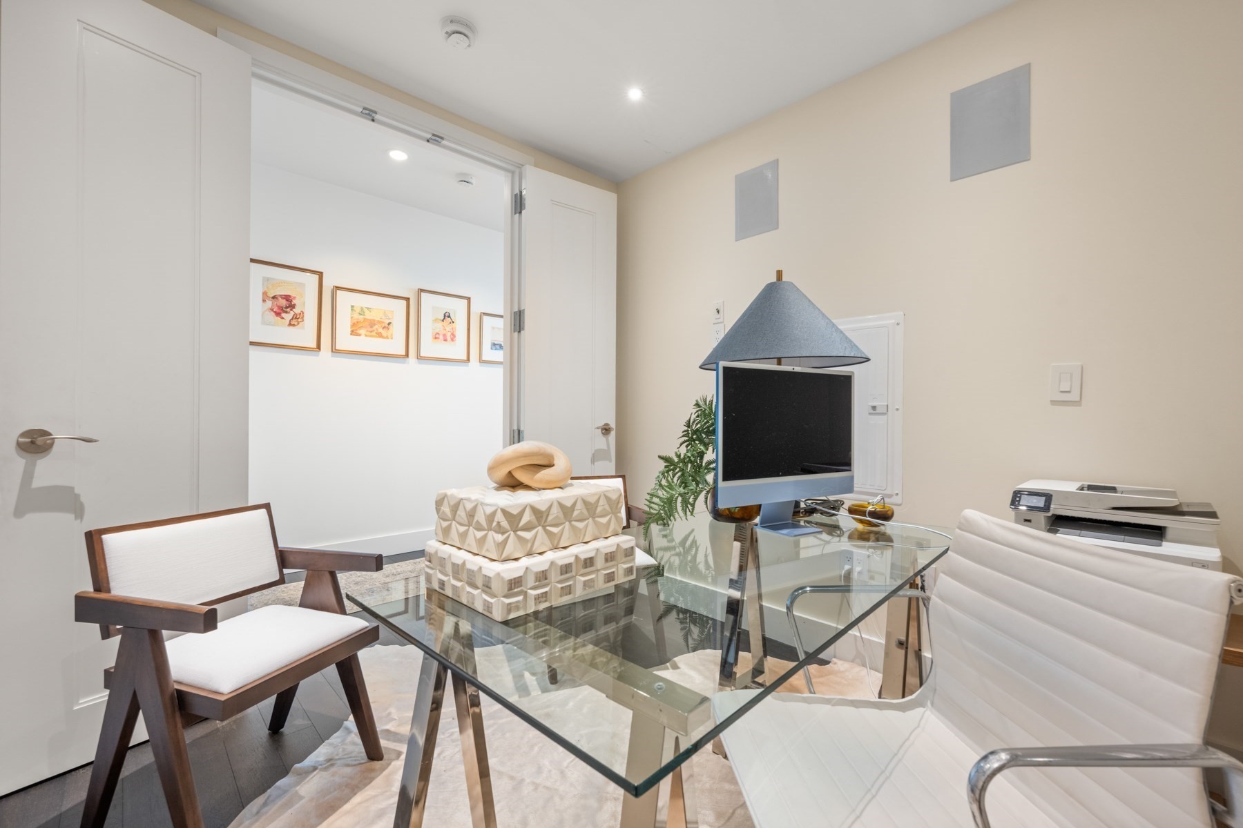 Furnished home office with a glass desk and white chairs, viewed through open double doors.
