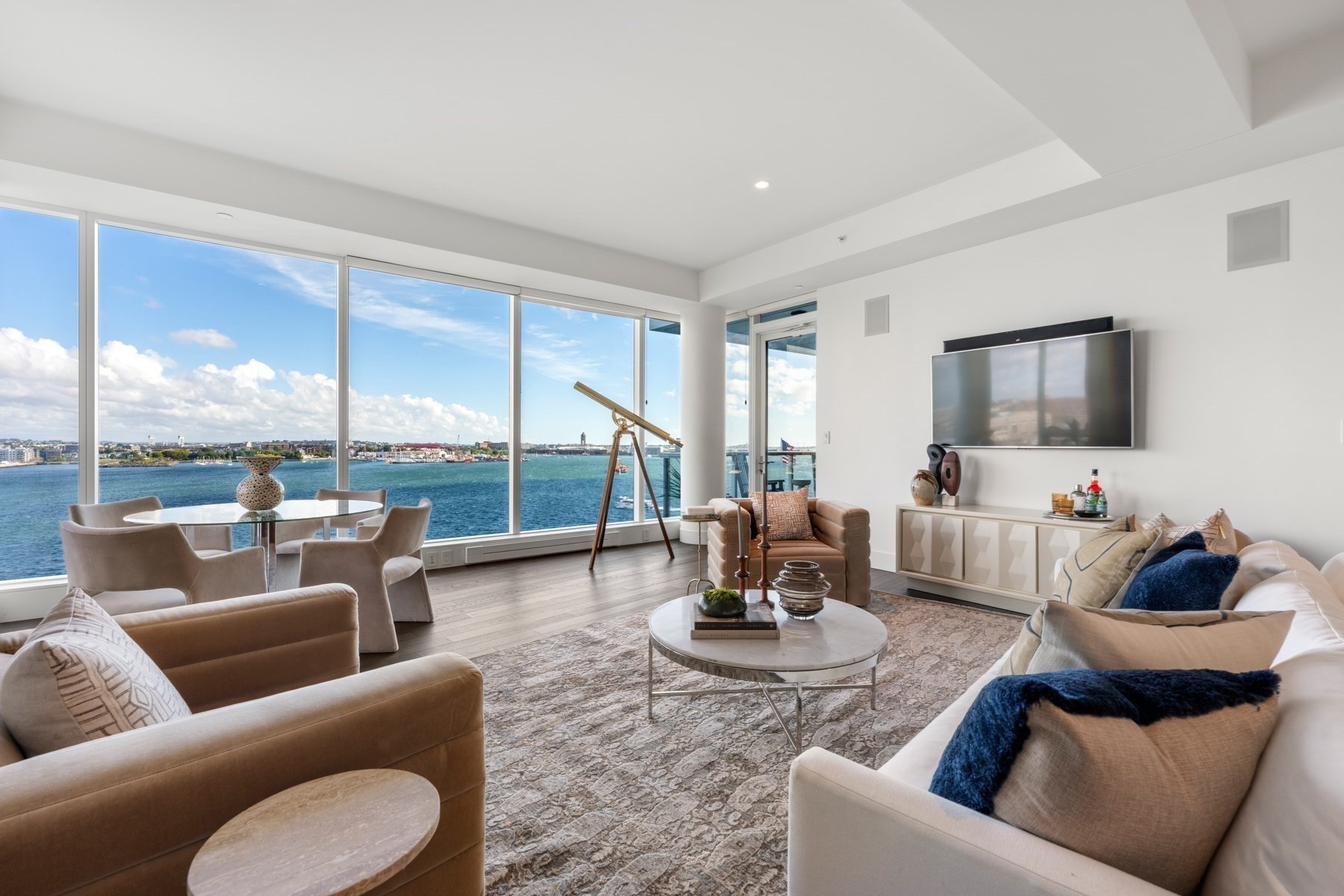Living room with floor-to-ceiling harbor views, neutral seating, and a telescope by the windows.