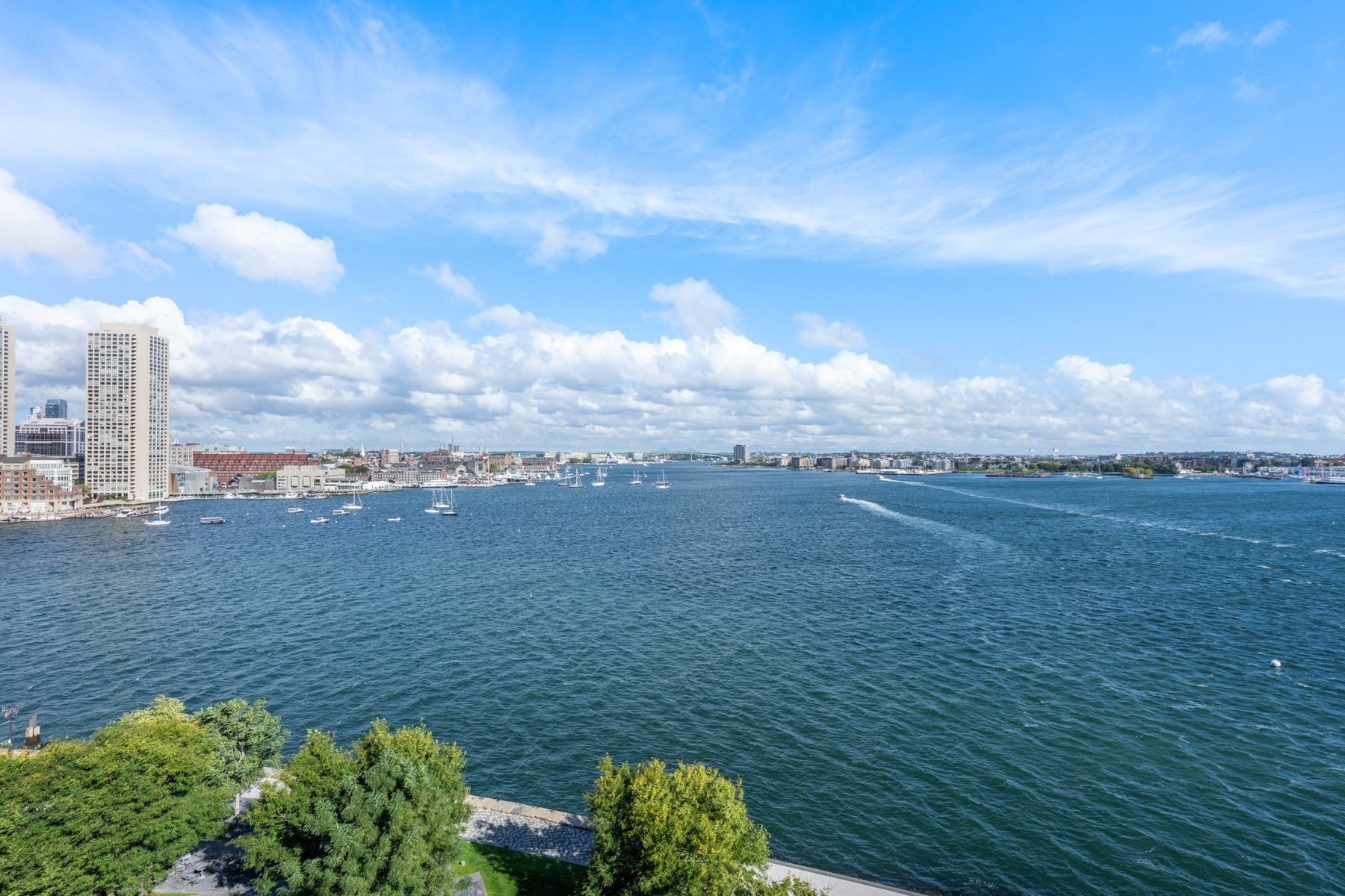 Boston Harbor view with sailboats, waterfront buildings, and a tree-lined promenade.