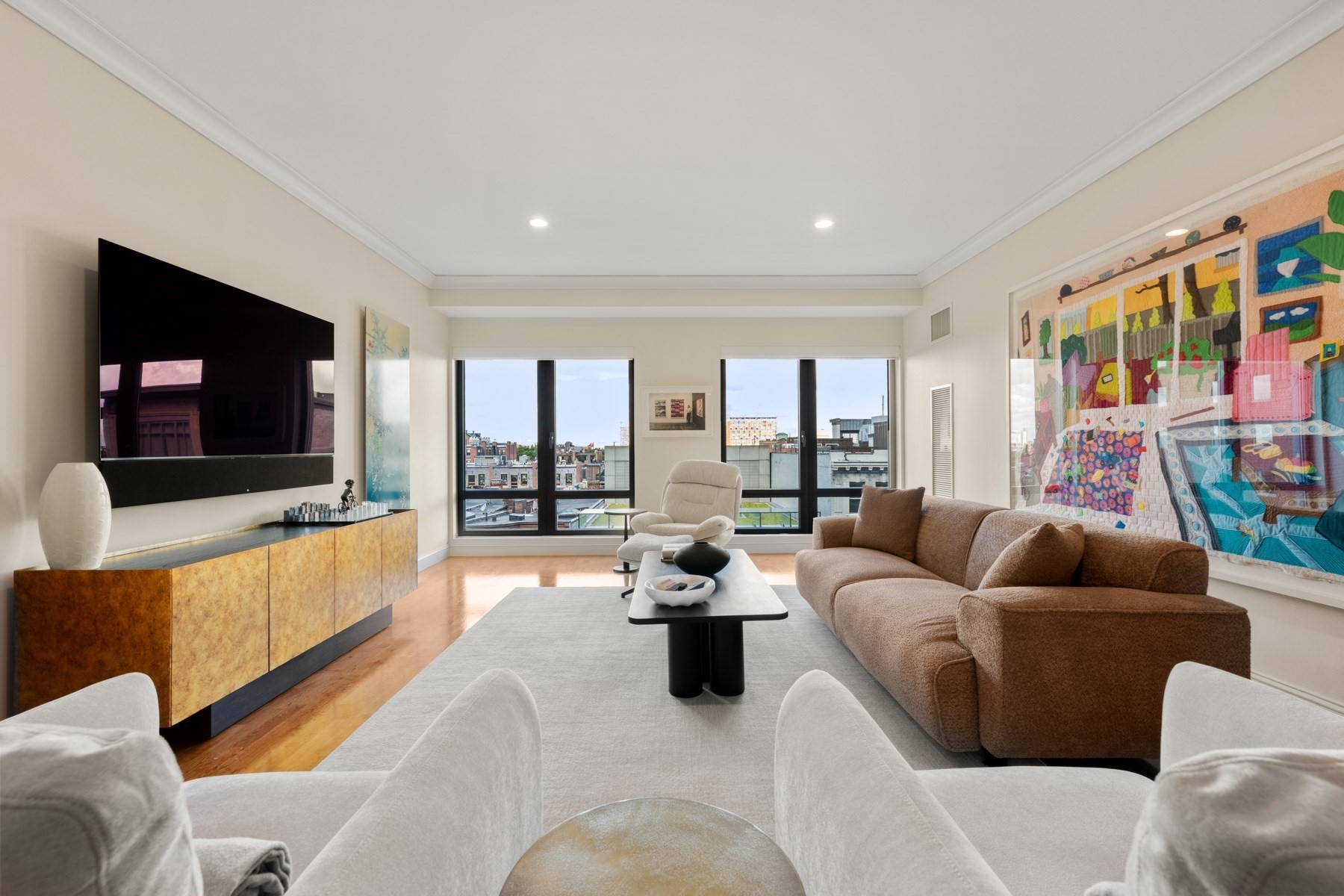 Living room with colorful wall art, a brown sectional, and large windows overlooking city rooftops.