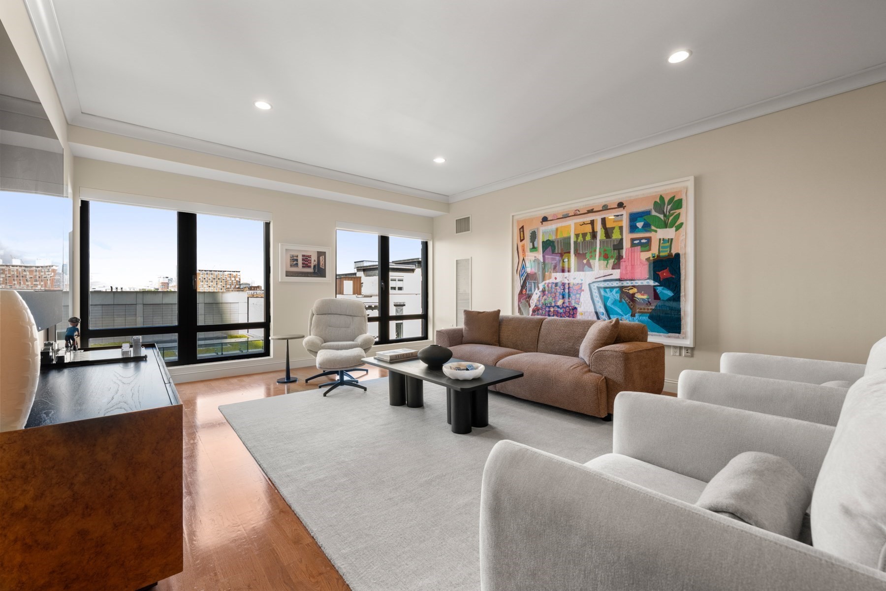 Living room with large windows, city views, a brown sofa, white armchairs, and colorful wall art.