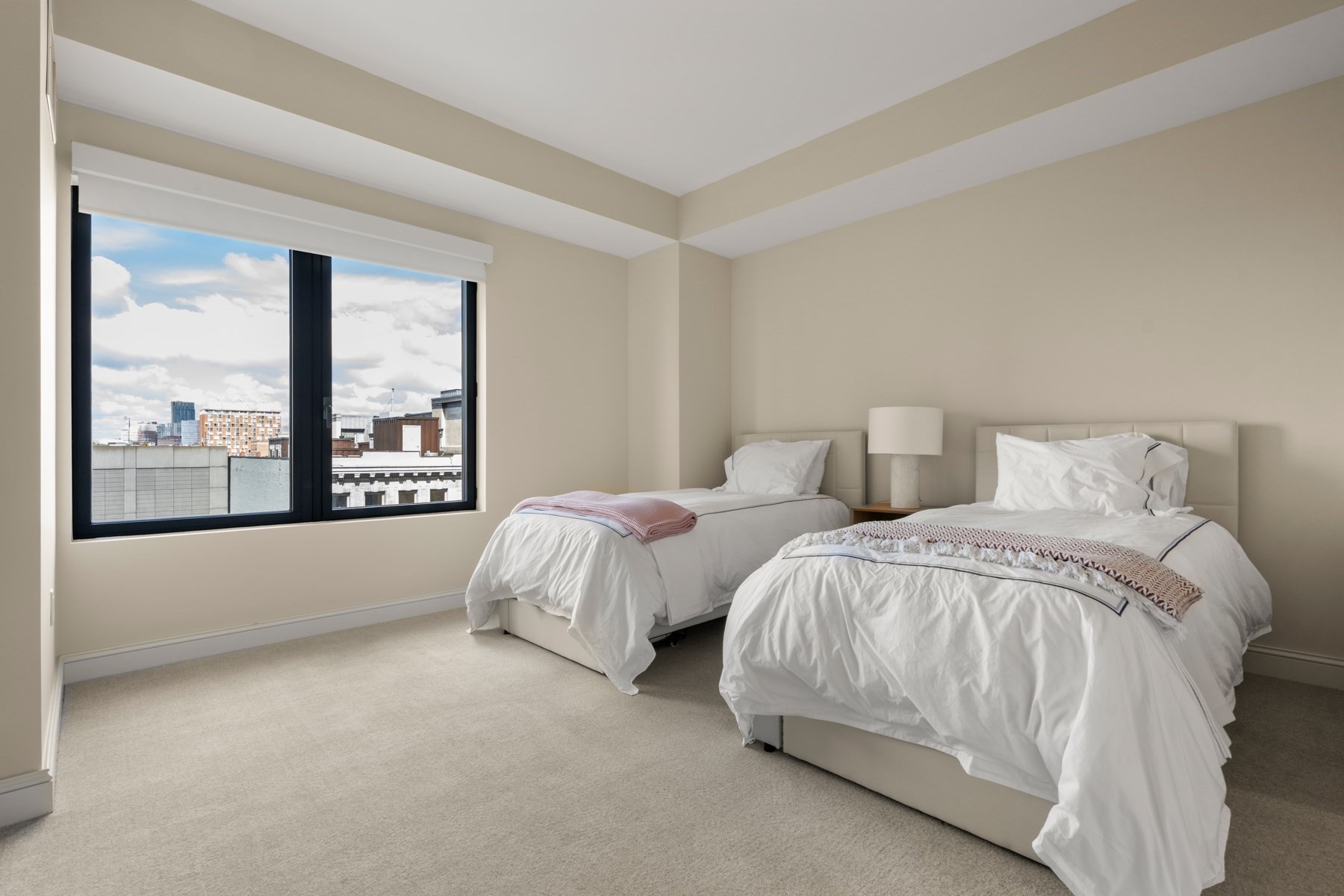 Bedroom with two white beds and a large window overlooking city buildings.
