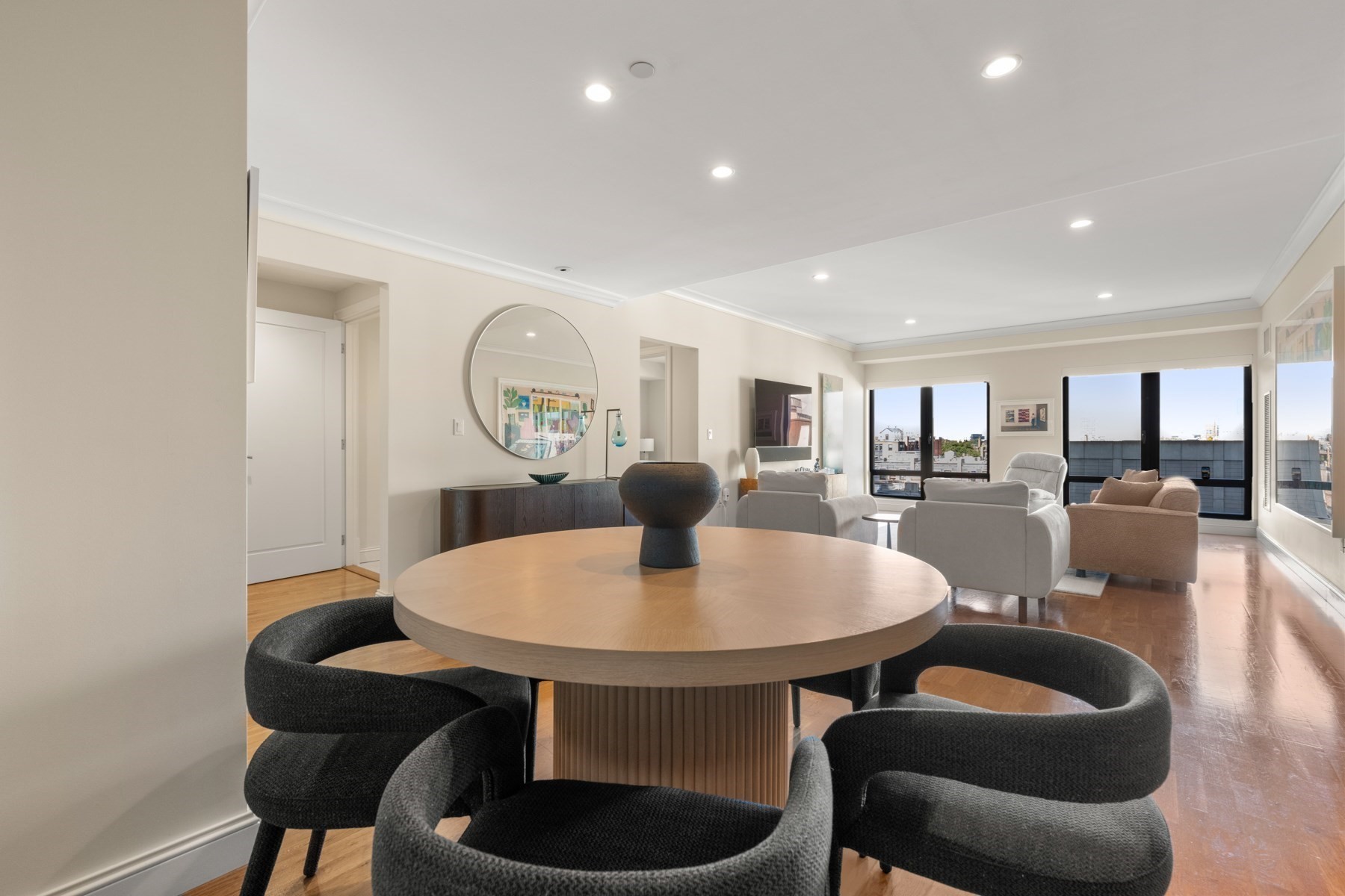 Open-plan dining and living room with a round table, upholstered chairs, and large city-view windows.