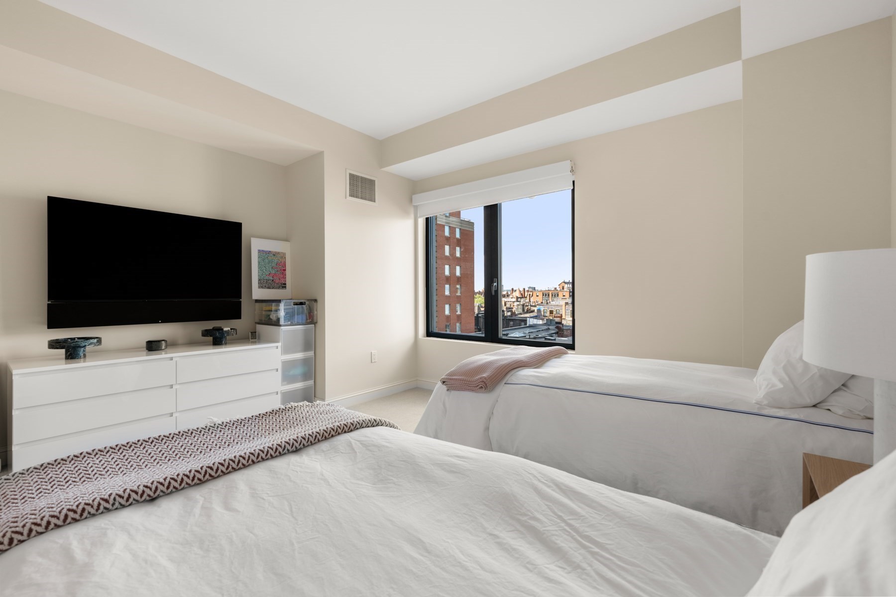 Furnished bedroom with two white beds and a large window overlooking city buildings.