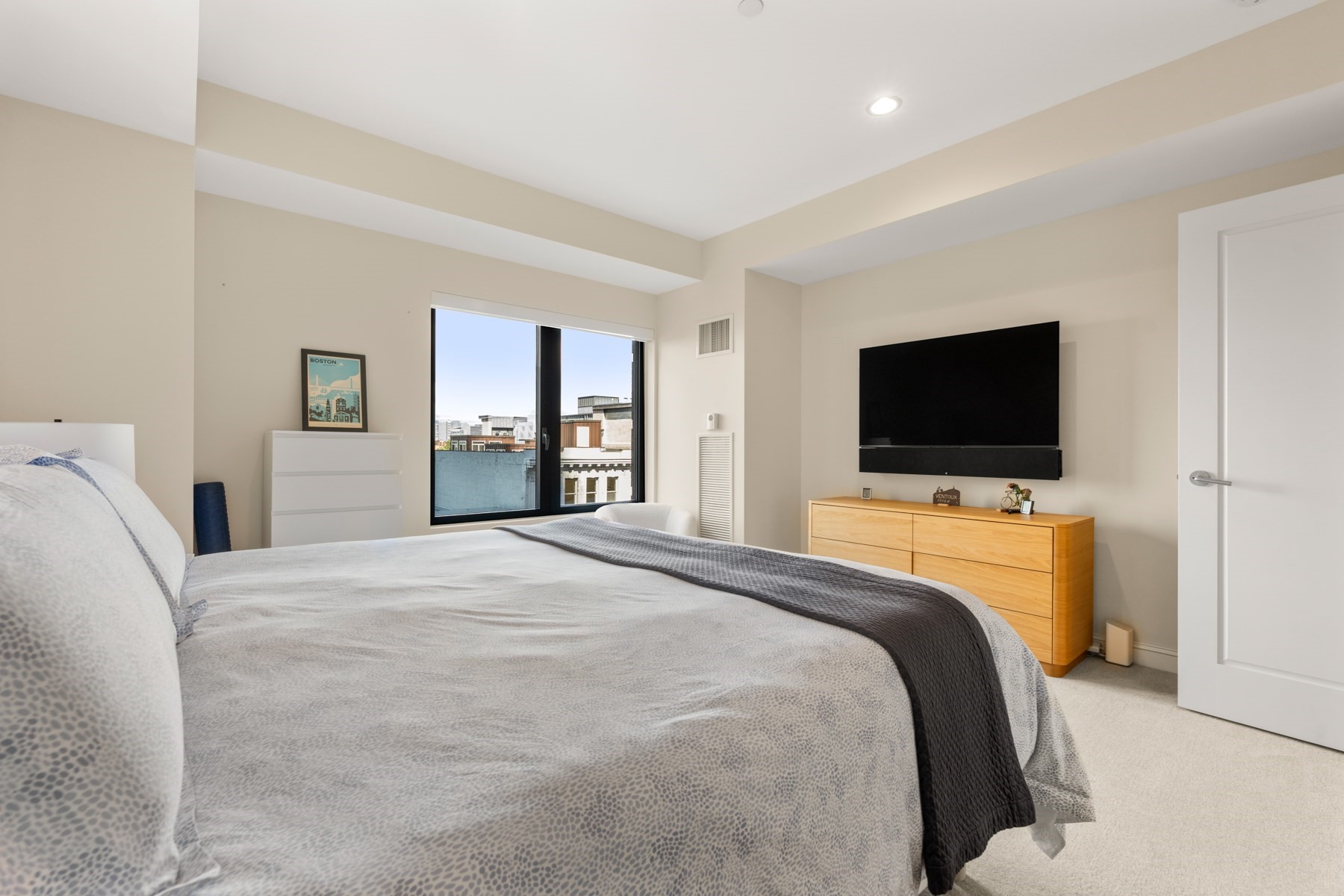 Bedroom with gray bedding, a wall-mounted television, and a window overlooking city buildings.