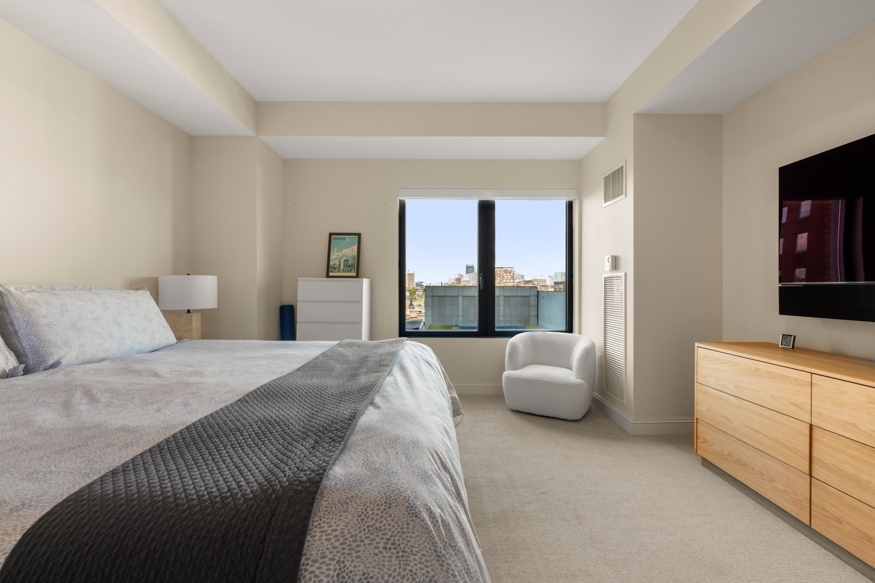 Bedroom with a large bed and armchair beside a window overlooking Boston buildings.