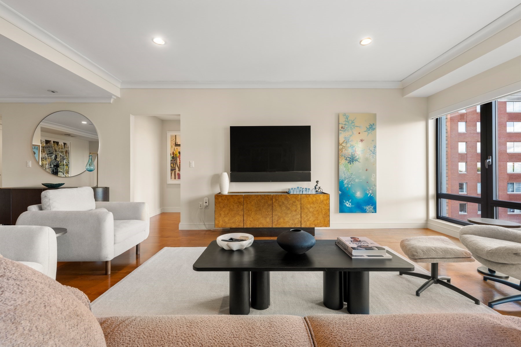 Living room with white seating, a wall-mounted TV, and large windows overlooking a brick building.