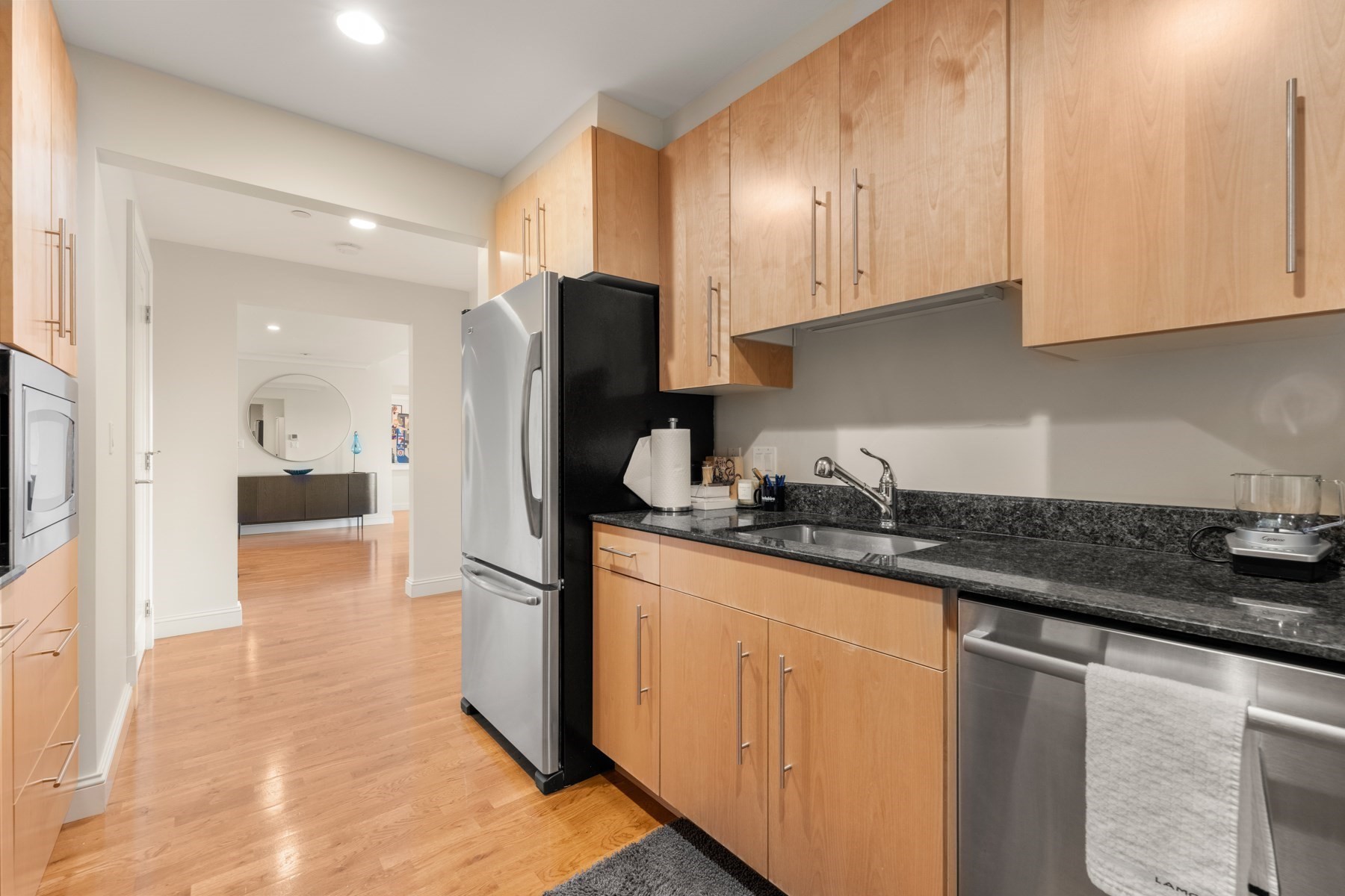 Condo kitchen with light wood cabinets, granite counters, and stainless-steel appliances opening onto a living area.