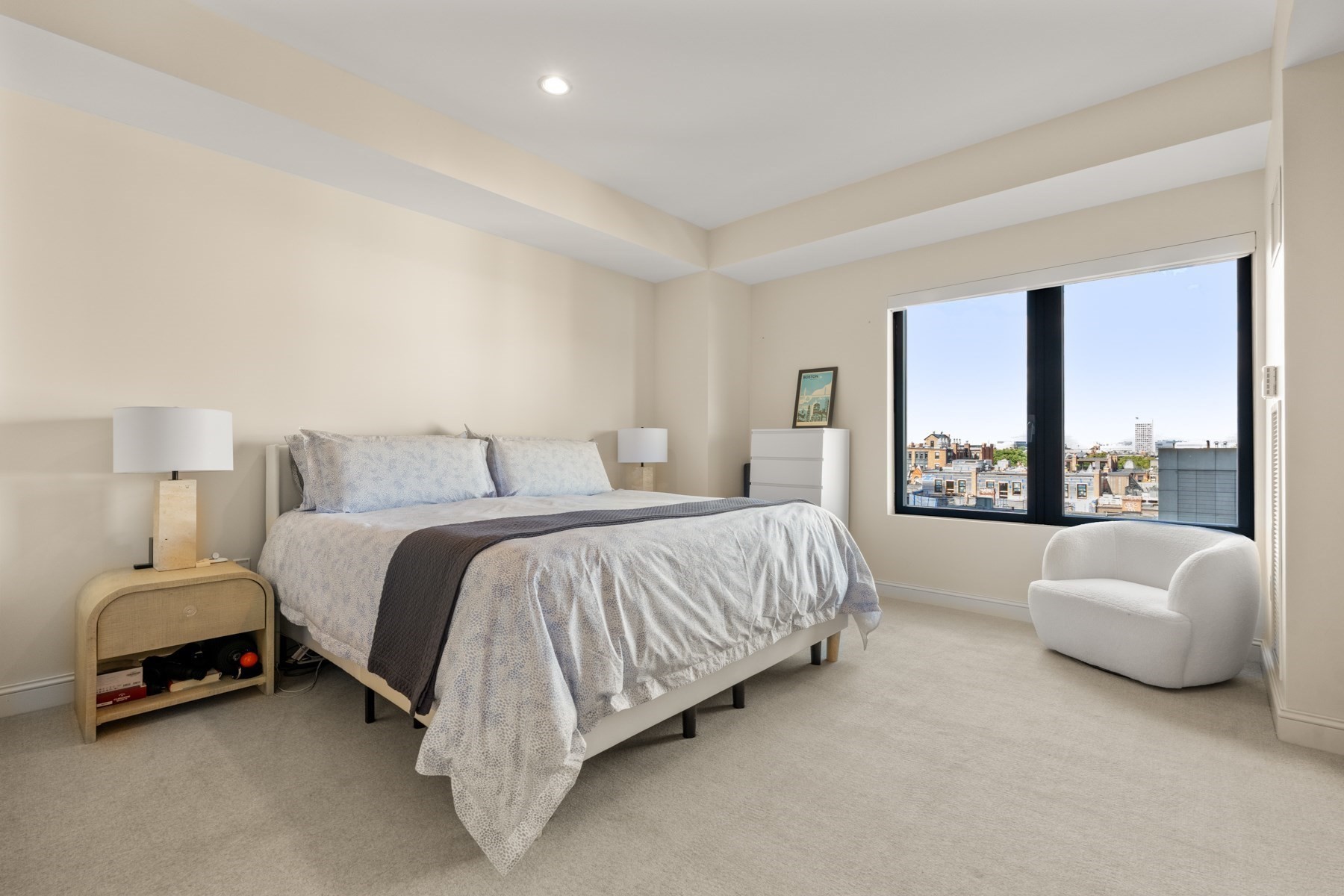 Staged bedroom with a large bed and white armchair beside a wide window overlooking city rooftops.