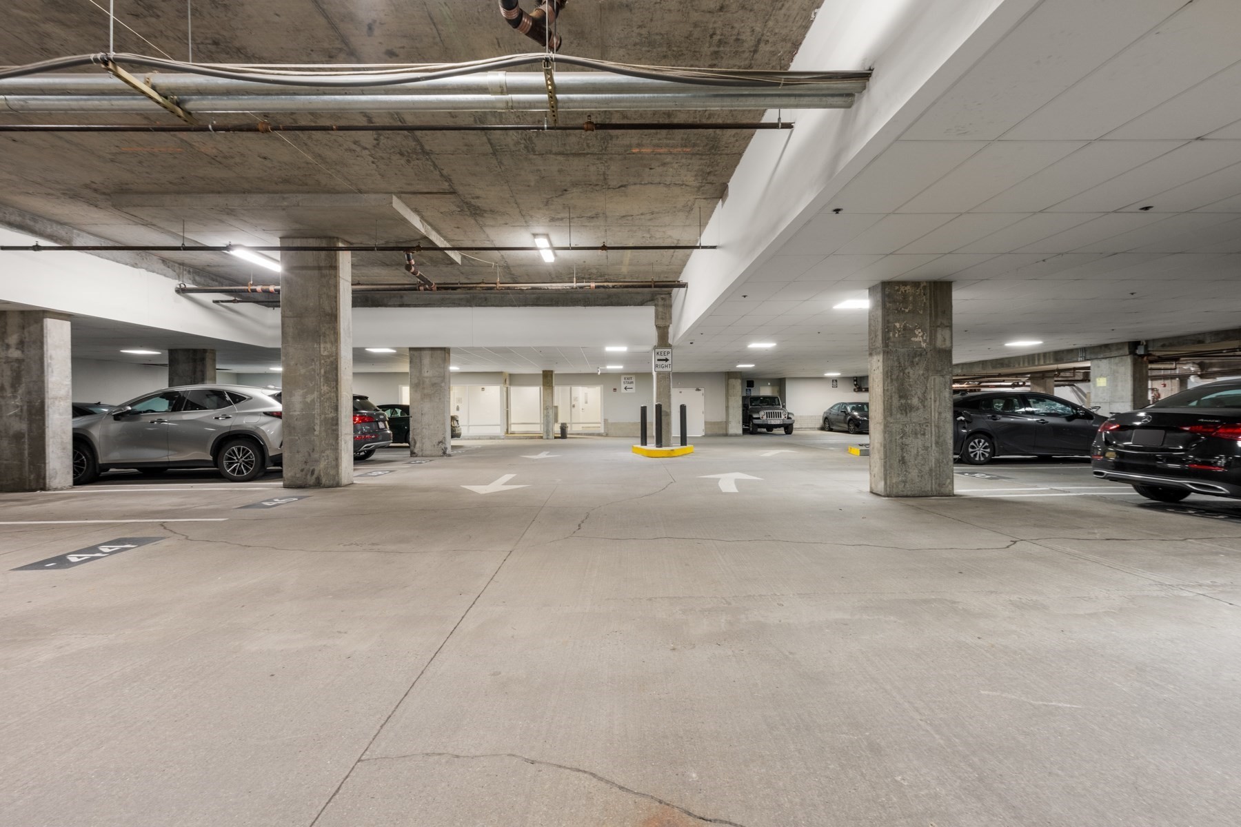 Underground parking garage with parked cars, concrete columns, and exposed ceiling pipes.