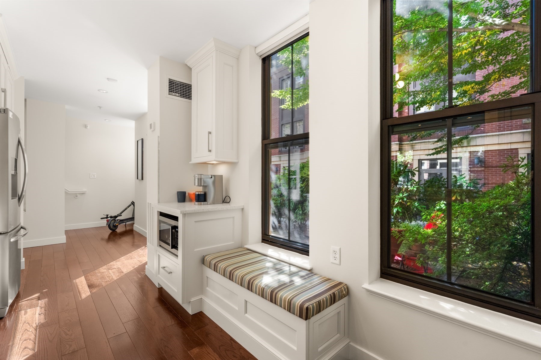 Bright kitchen with white cabinetry, a built-in coffee counter, and a striped window seat beside leafy views.