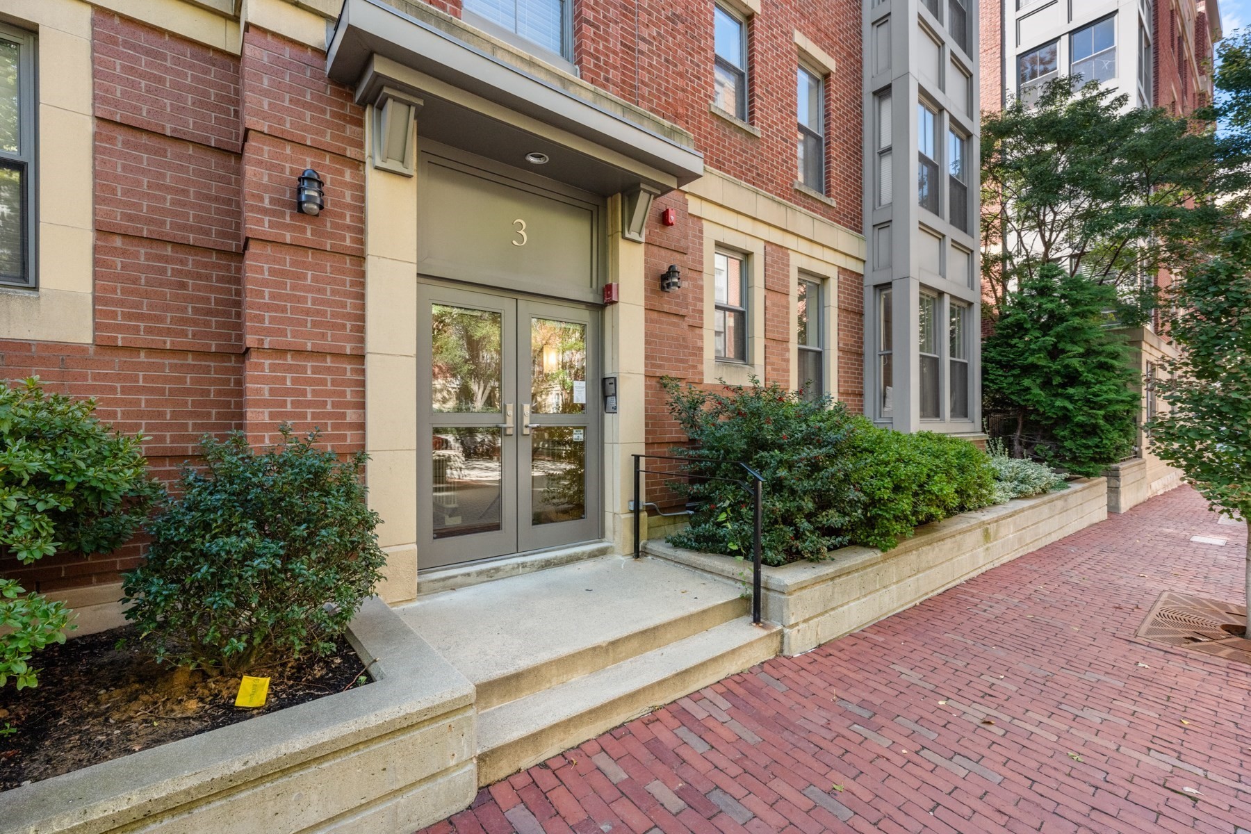 Brick residential building entrance with glass doors, landscaped planters, and a brick walkway.