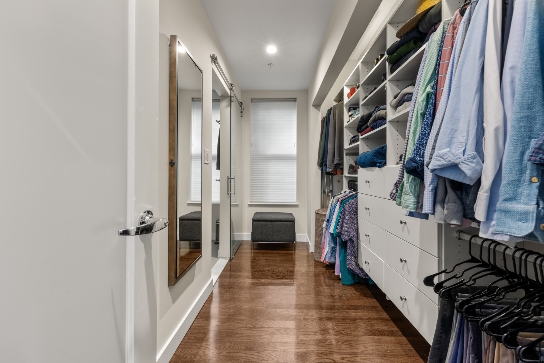 Walk-in closet with built-in shelves and drawers, hanging clothes, and a bench beneath the windows.