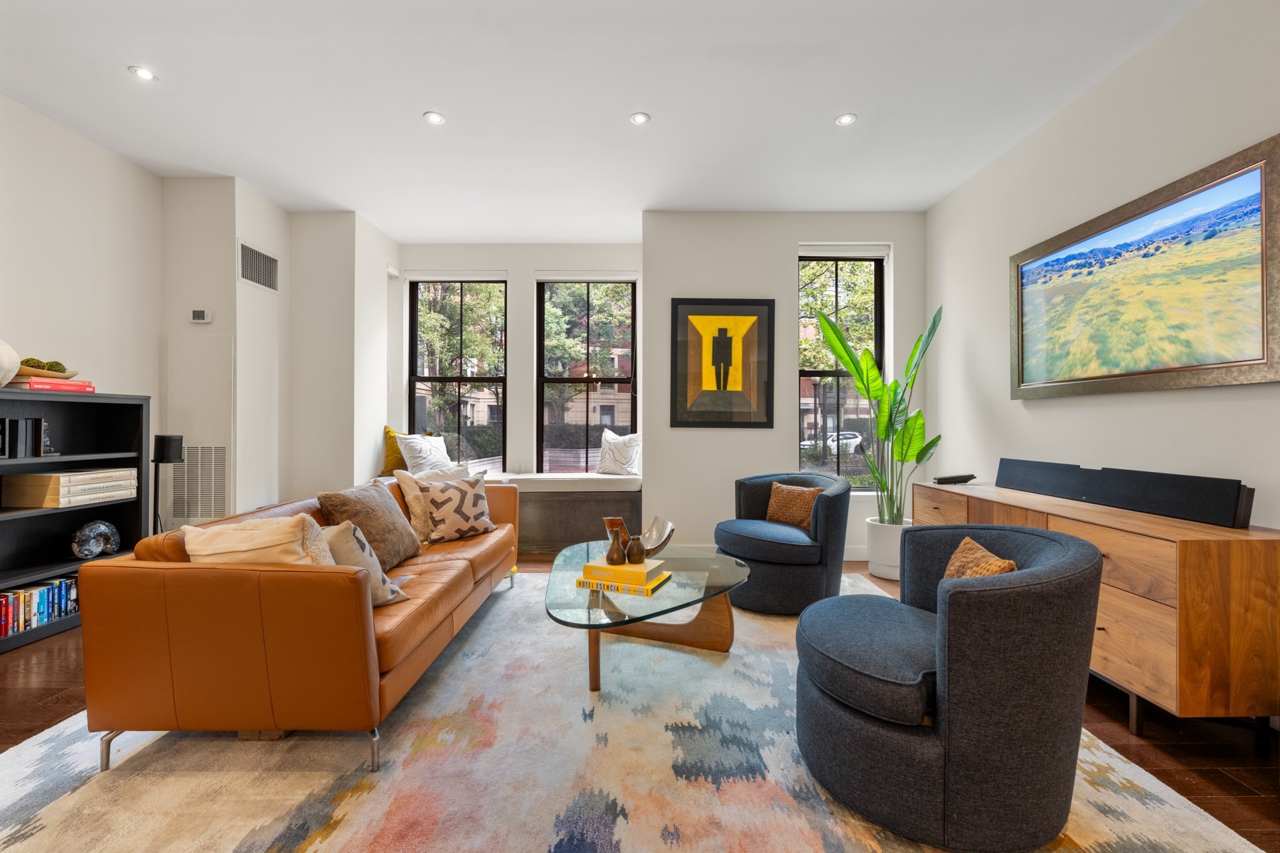Furnished living room with a caramel leather sofa, charcoal chairs, and large windows overlooking trees.