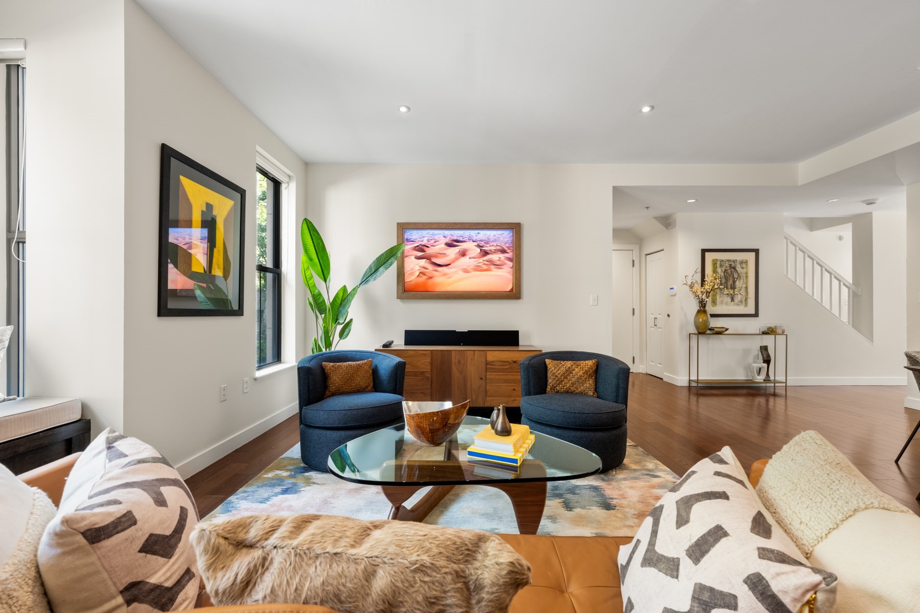 Living room with blue accent chairs, hardwood floors, colorful rug, and an open staircase in the background.
