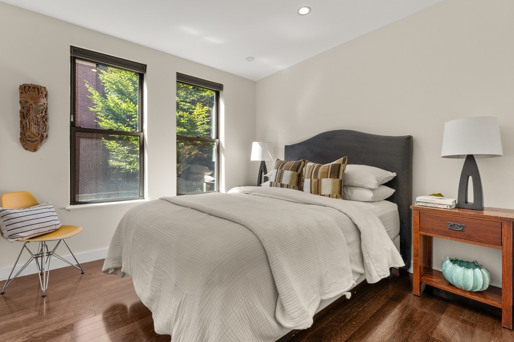 Bedroom with a charcoal headboard, hardwood floors, and tall windows overlooking leafy greenery.