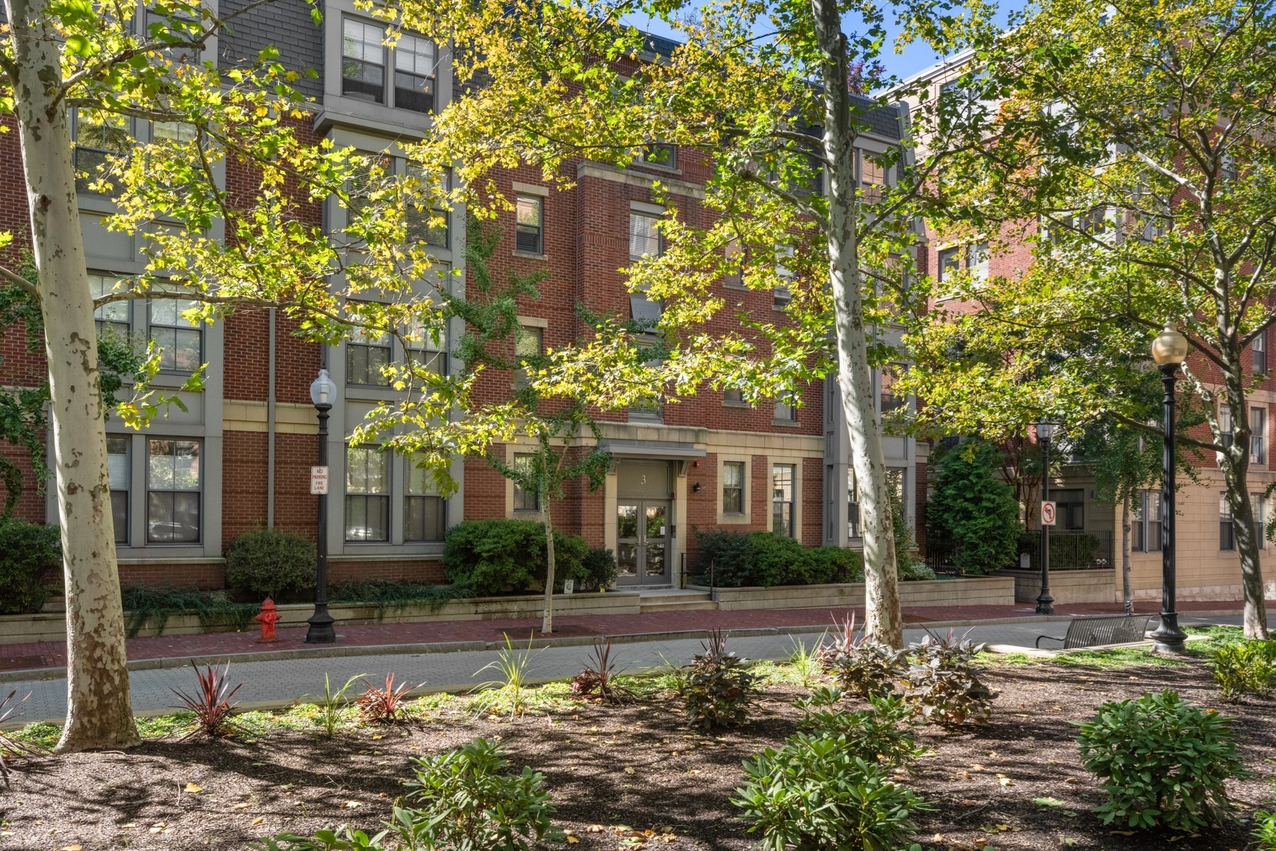 Red-brick residential building with a central entrance, framed by leafy trees and landscaped planting beds.