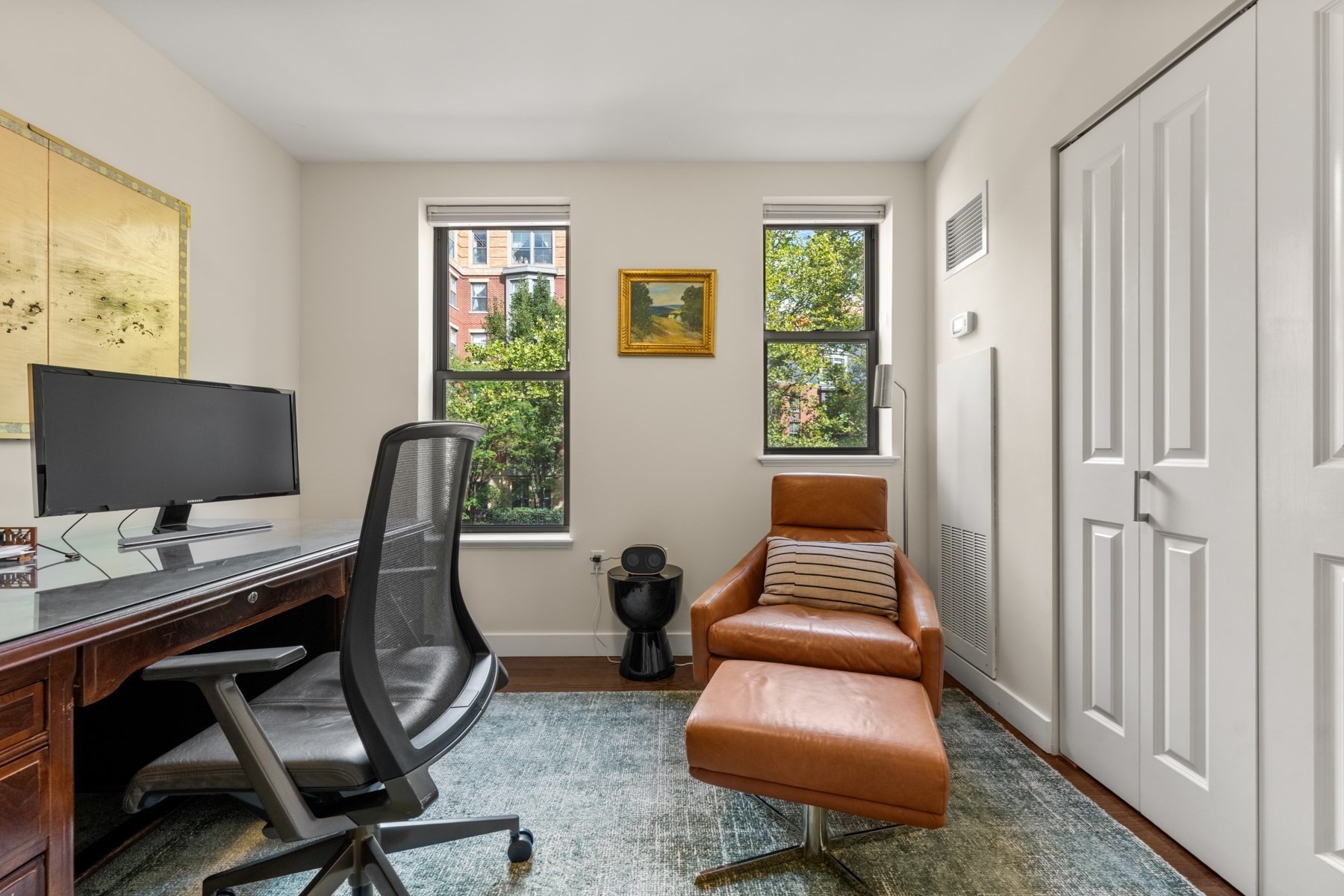 Furnished home office with a wood desk, two windows, and a brown leather lounge chair and ottoman.