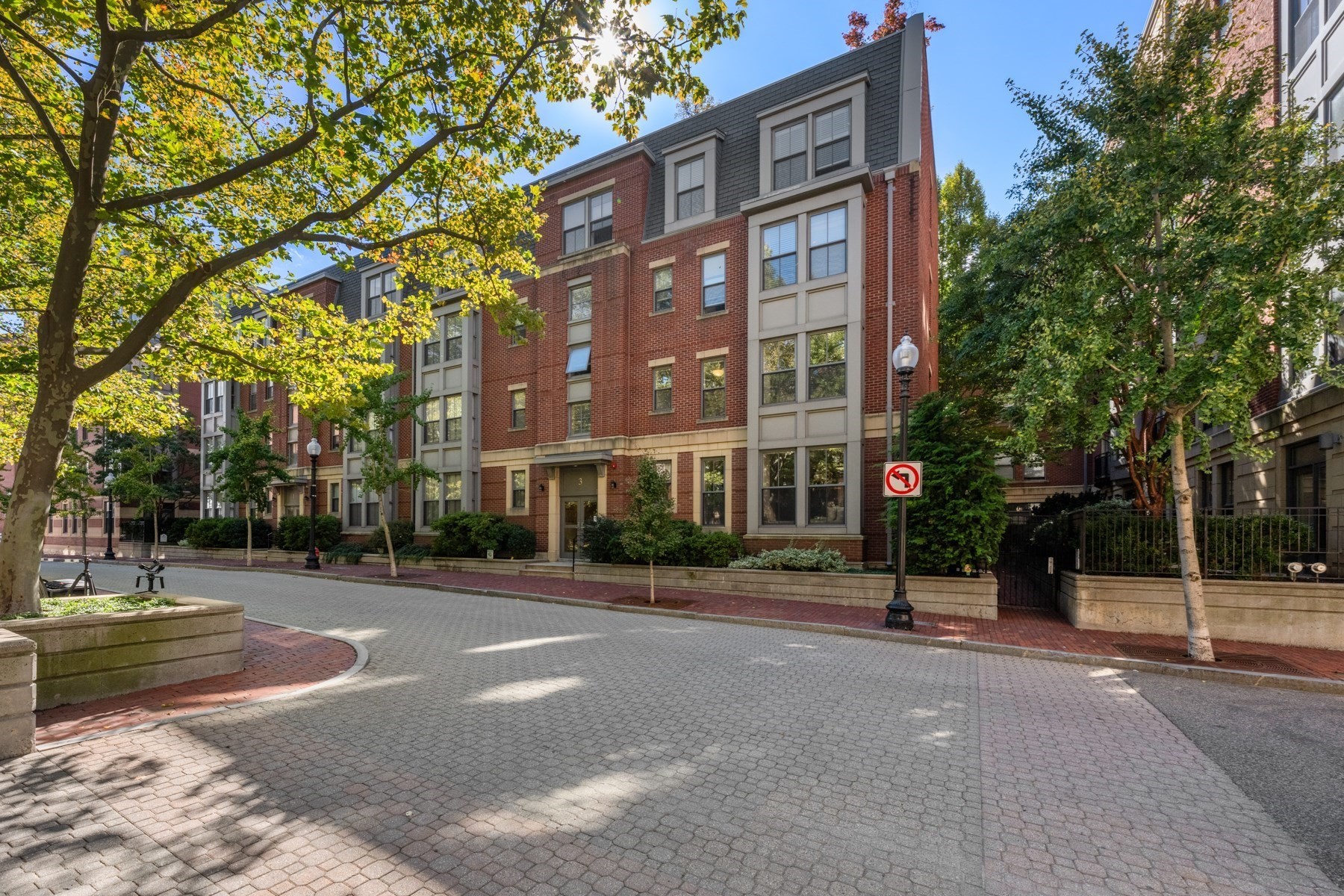 Red-brick residential building with gray dormers, mature trees, and a cobblestone street.