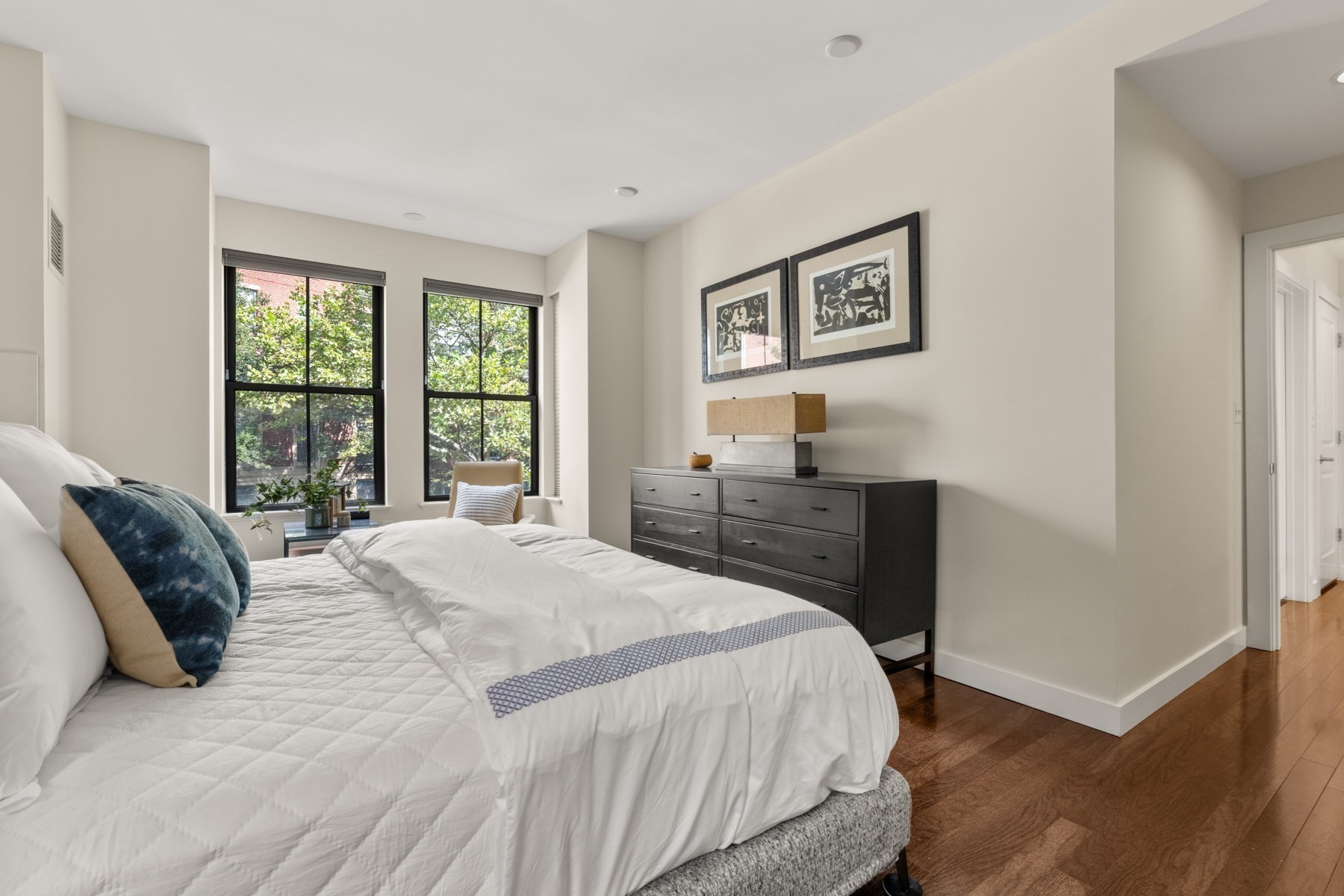 Staged bedroom with a white quilted bed, dark dresser, and large windows overlooking trees.