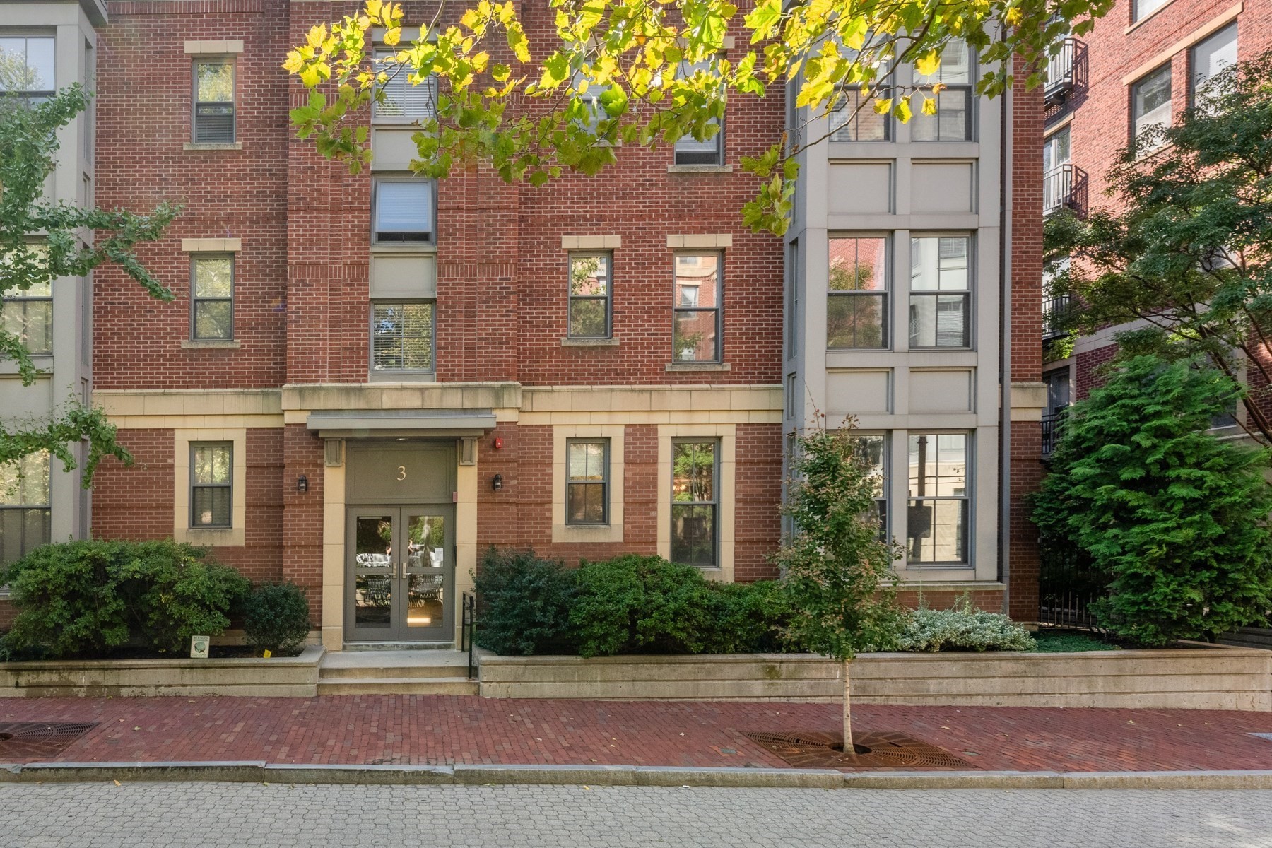 Red-brick residential building entrance marked 3, framed by shrubs and a brick sidewalk.