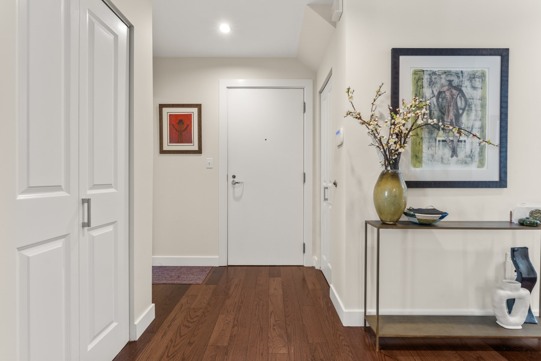 Apartment entry hall with dark hardwood floors, framed art, and a console table with flowering branches.