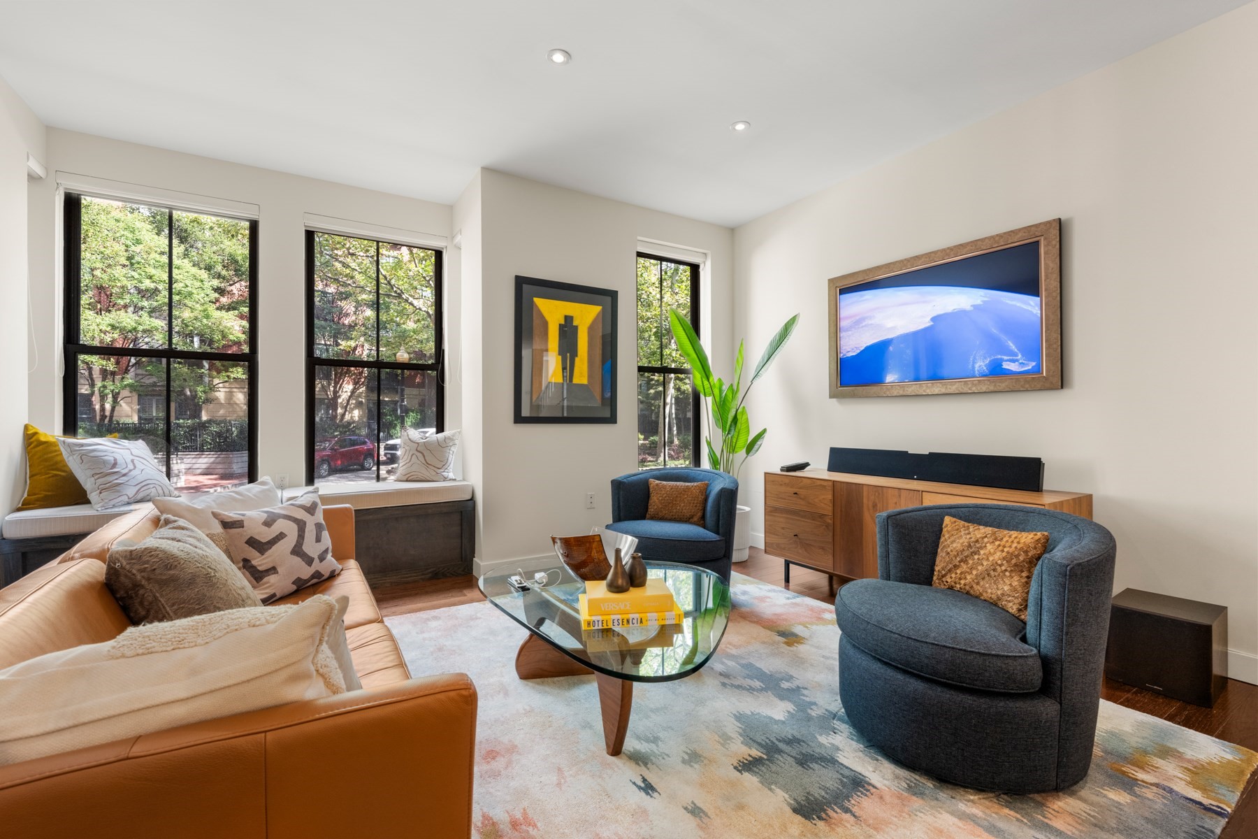 Living room with a tan leather sofa, blue armchairs, and tall windows overlooking leafy trees.