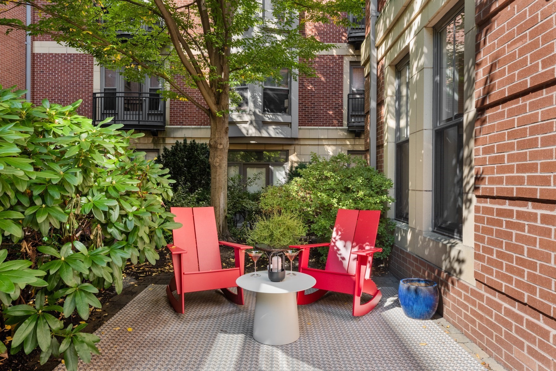 Courtyard patio with red Adirondack chairs, a white table, mature trees, and brick apartment buildings.