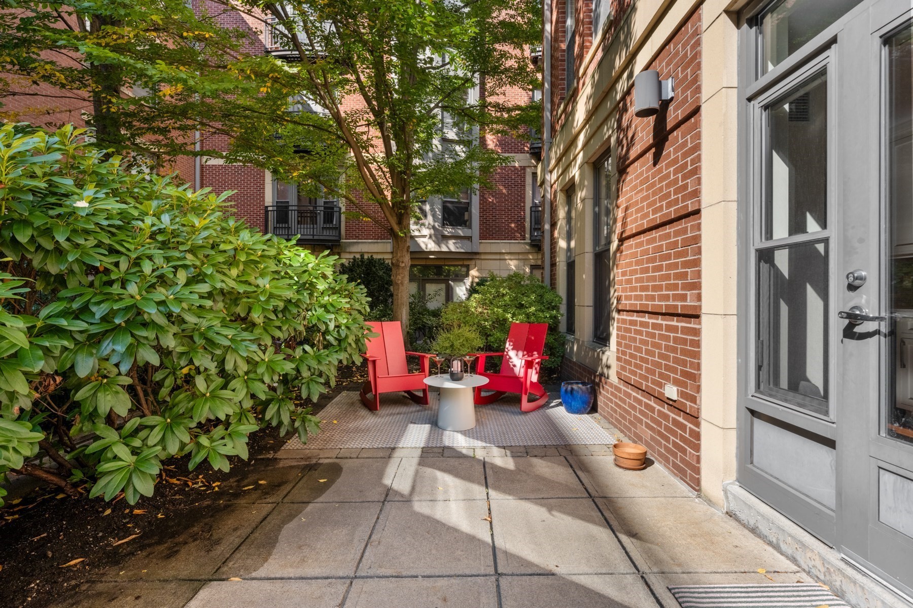 Courtyard patio with red chairs, a small table, and leafy shrubs beside a brick building.