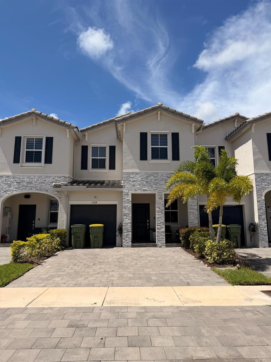 Attached two-story townhomes with stone veneer, dark shutters, and a palm beside the paved driveway.