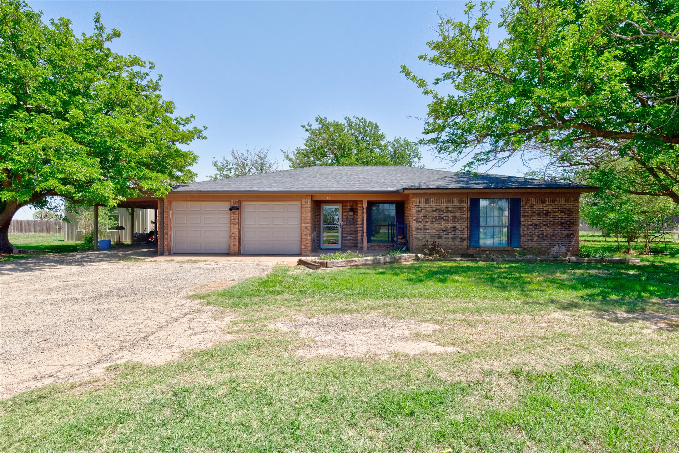 Single-story brick house with a two-car garage and carport, framed by mature trees and a broad lawn.