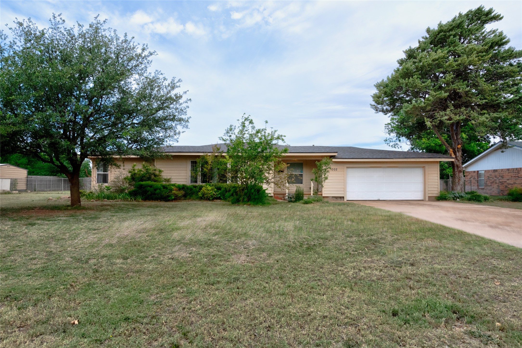 Single-story beige house with a white double garage, broad lawn, and mature trees.
