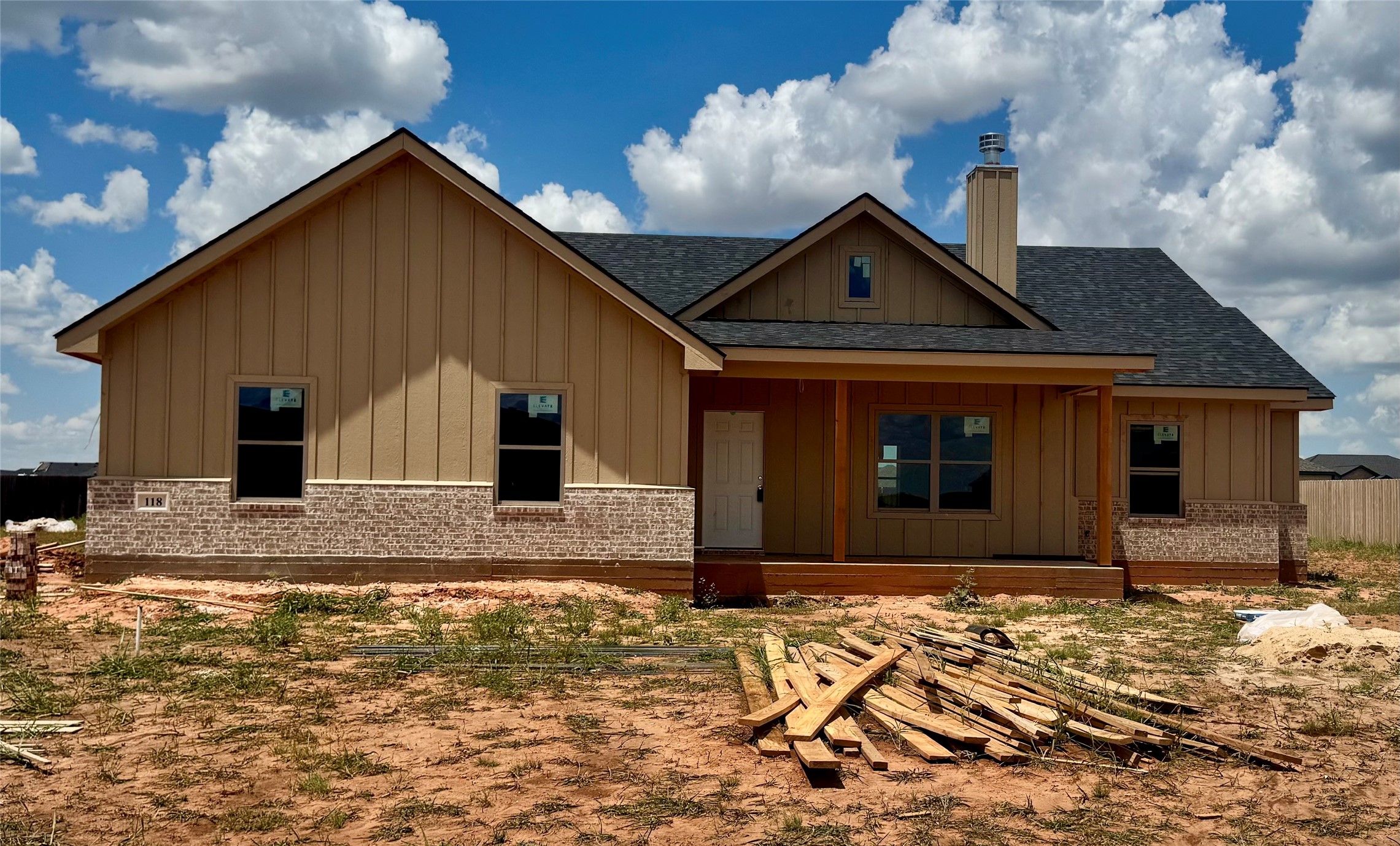 Tan house under construction with a brick lower façade, covered porch, and lumber scattered across the yard.