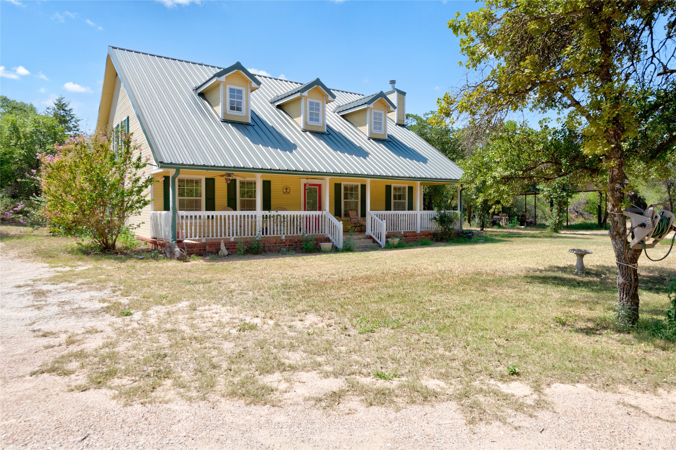 Yellow home with a steep metal roof and covered porch in a tree-shaded yard.